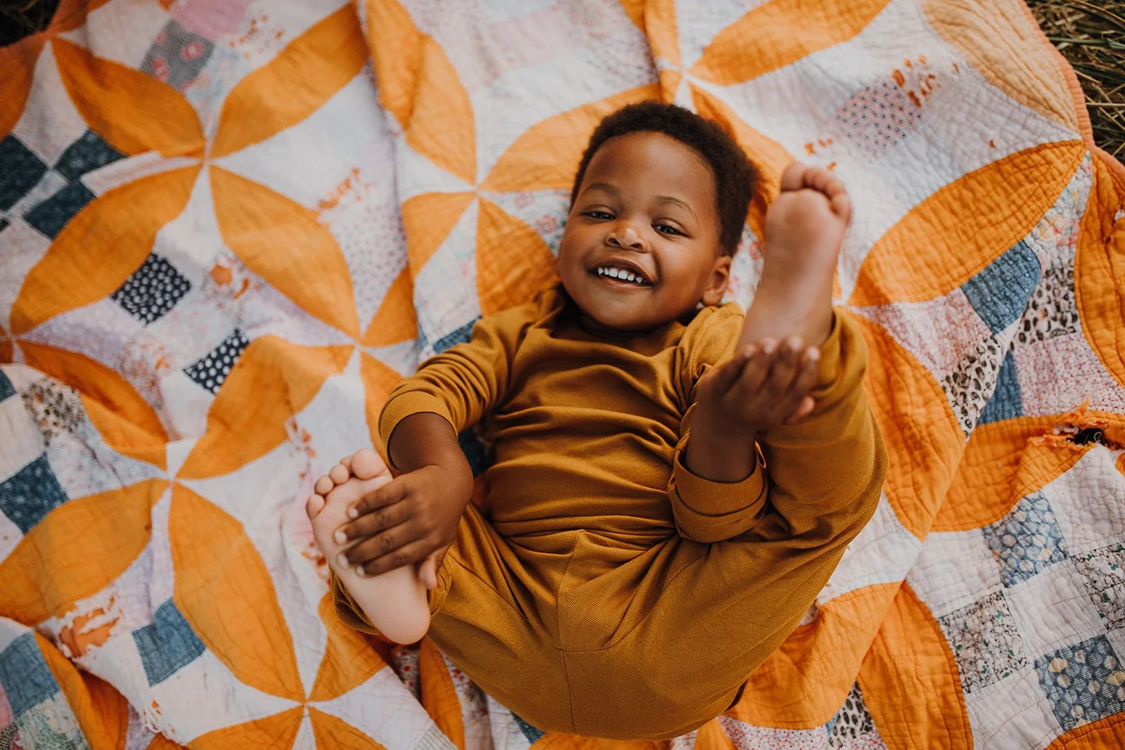 A smiling young child lying on a colorful quilt, playing with their foot, dressed in a brown outfit.