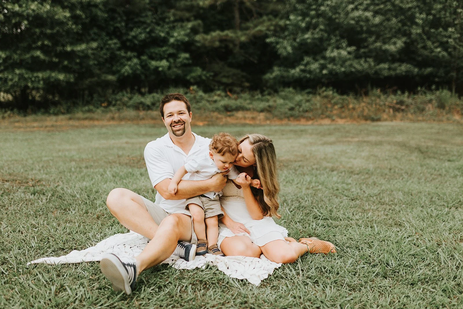 Family of three sitting on a blanket in a grassy field, smiling and playing together. The father, mother, and young son are enjoying a sunny day outdoors with trees in the background.
