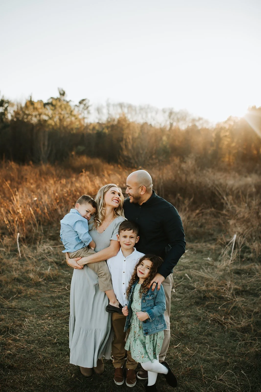 A family of six celebrating outdoors during sunset, with autumn trees in the background. The mother and father are smiling at each other, with the children surrounding them, all appearing happy and affectionate.