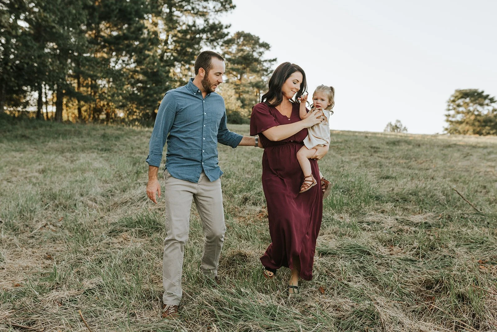 A family of three walking outdoors in a grassy field with trees in the background during sunset, the mother carrying a young girl and the father walking beside them.