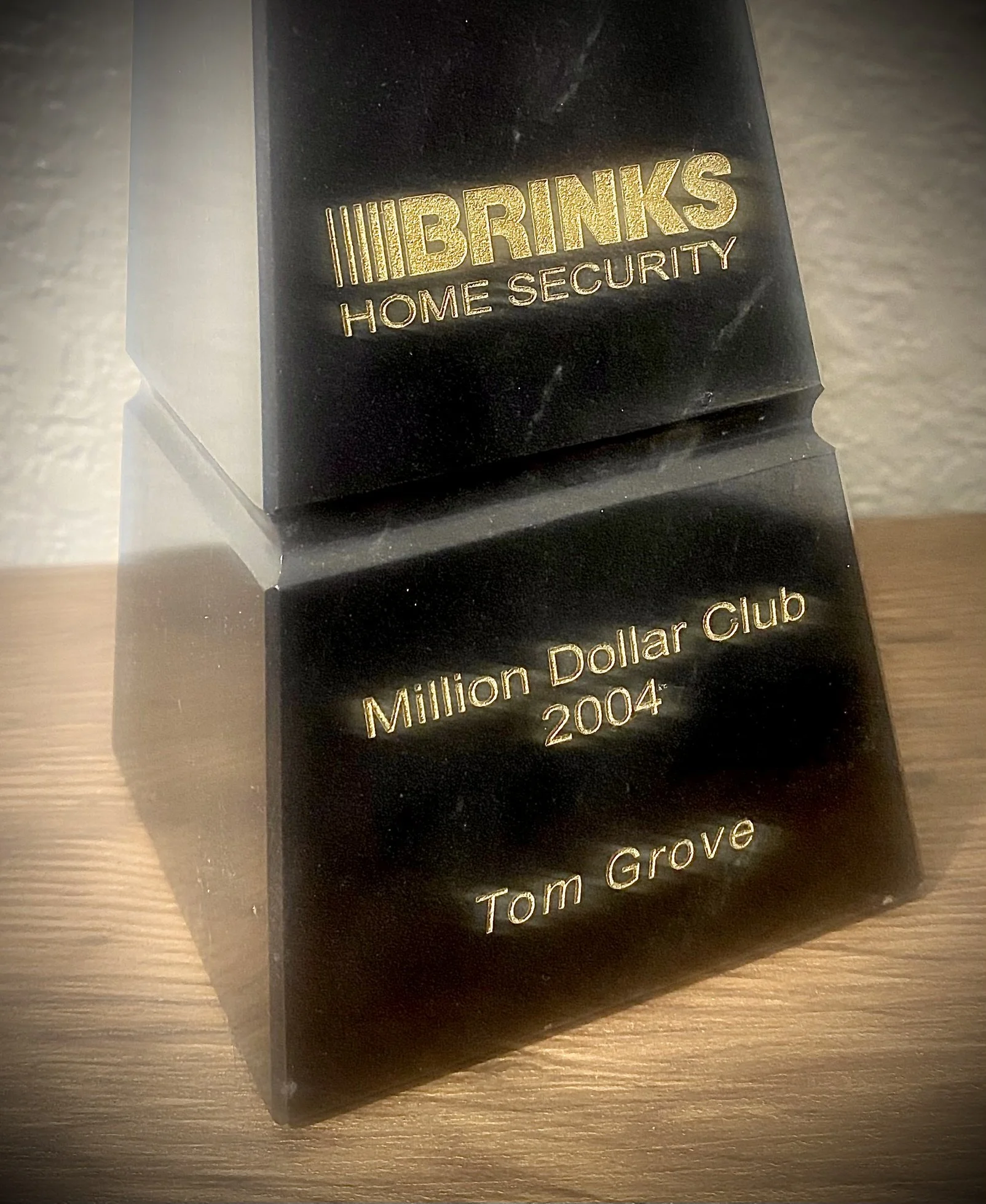 Black trophy with gold engraving reading 'BRINKS HOME SECURITY', 'Million Dollar Club 2004', and 'Tom Grove', placed on a wooden surface.