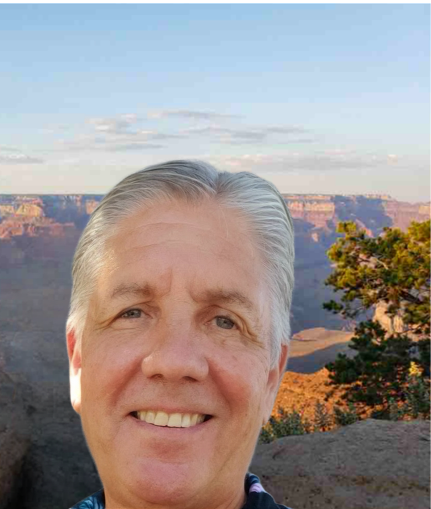 A smiling man with gray hair taking a selfie at the Grand Canyon during sunset or sunrise, with the canyon and a partly cloudy sky in the background.