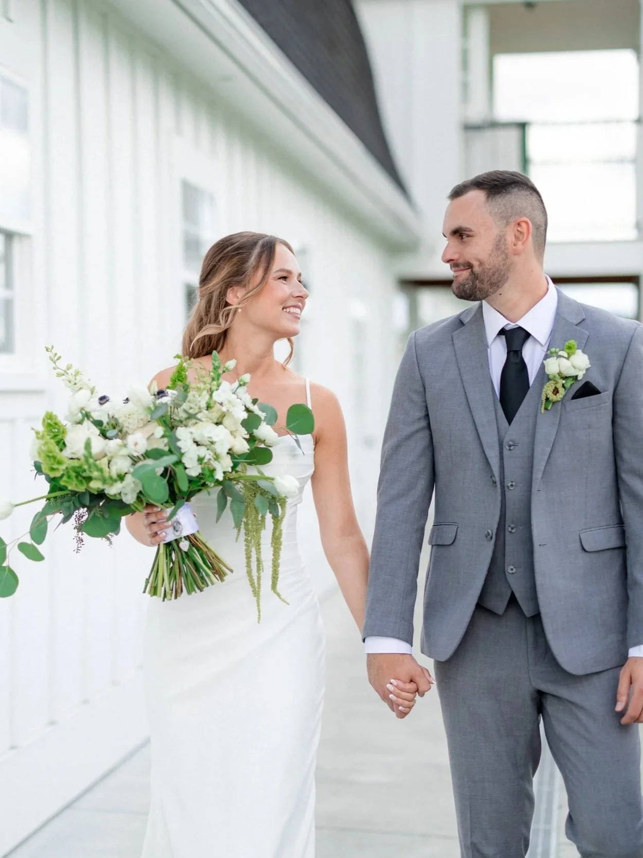 Bride and groom walking hand-in-hand outside, smiling at each other, bride holding a bouquet of white and green flowers, groom in a gray suit with boutonniere.