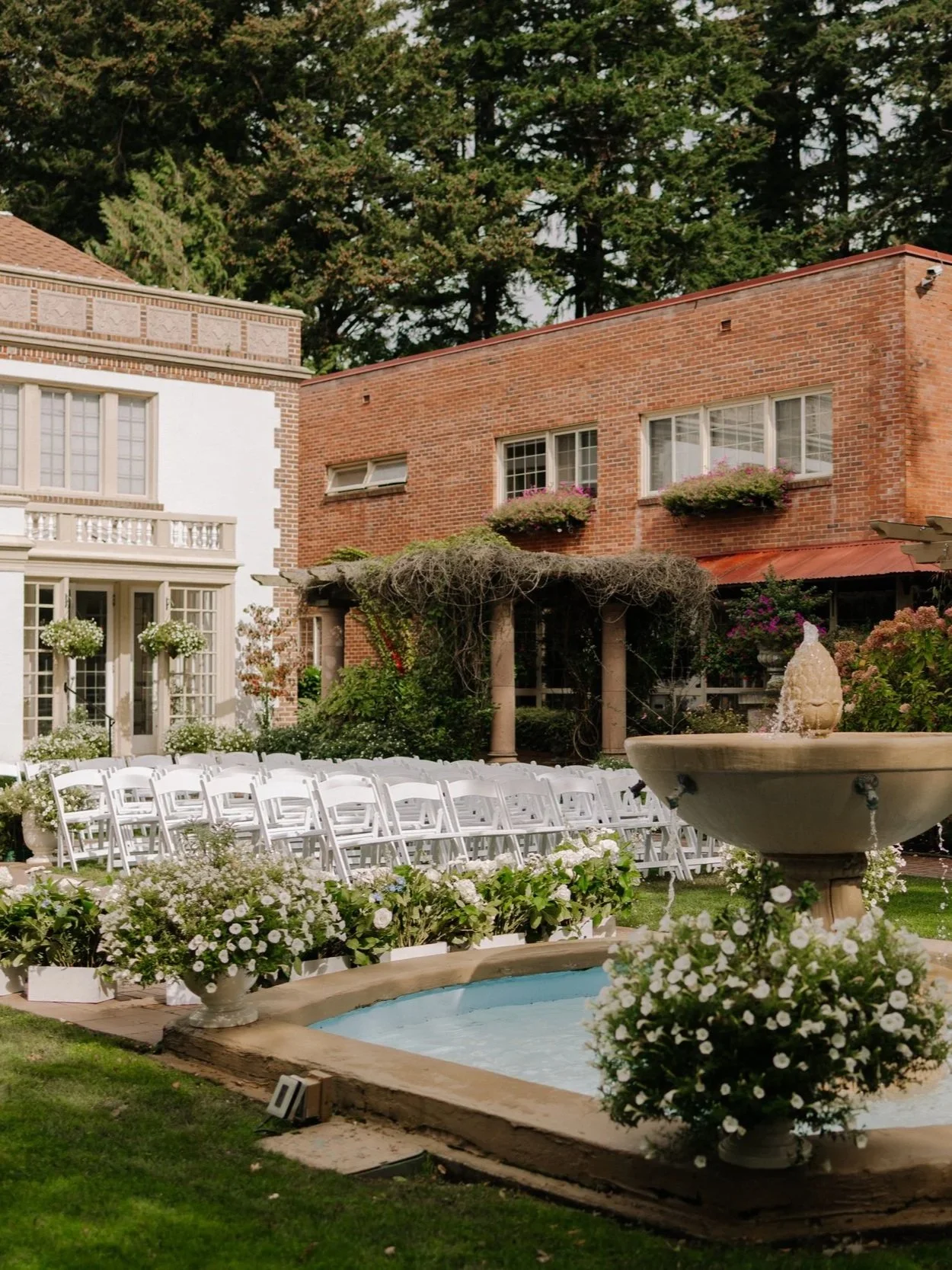 A garden courtyard with a fountain surrounded by white flowers, white chairs arranged in rows, and a pergola, in front of historic buildings with brick and white facades, and trees in the background.