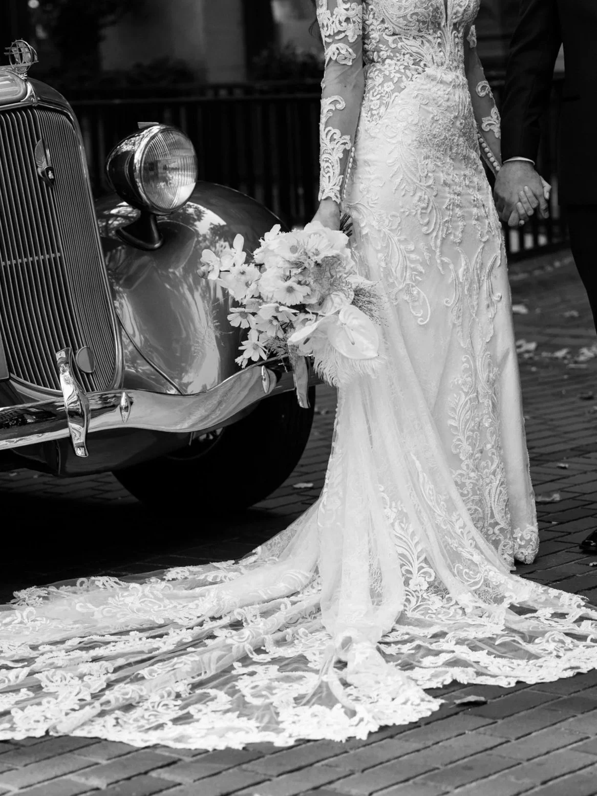 A bride and groom holding hands next to a vintage car with a lace wedding dress train and a bouquet of flowers.