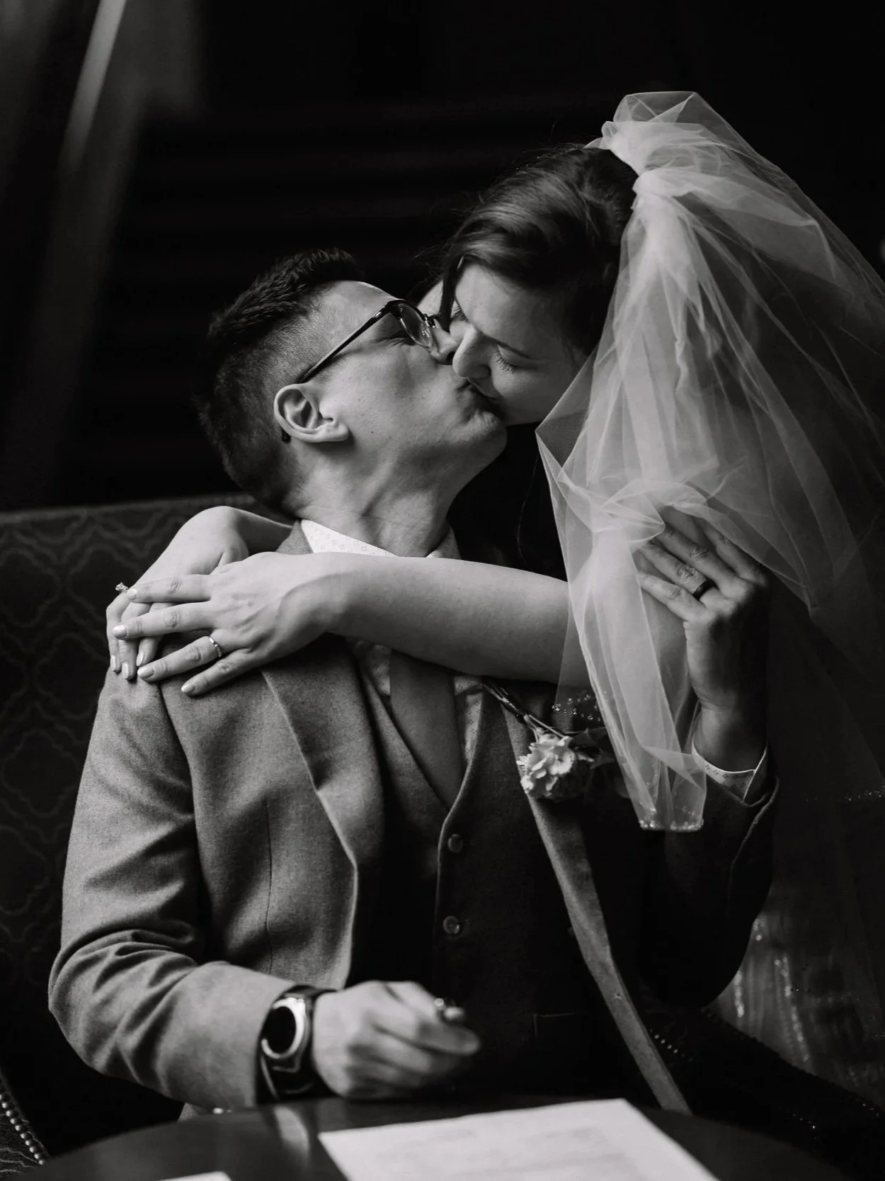 A black and white photo of a bride and groom sharing a kiss. The bride is leaning over the groom, who is seated, and they are embracing.