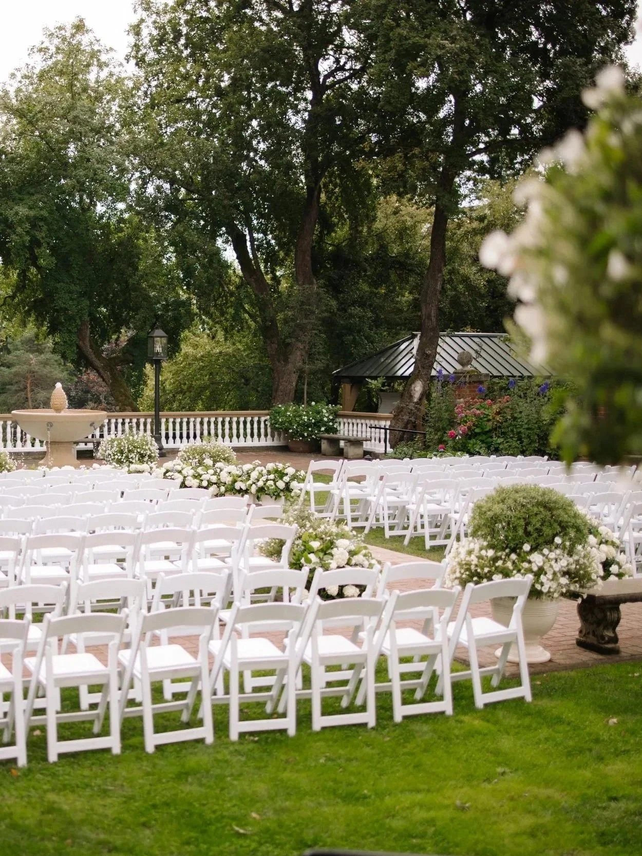 Outdoor wedding setup with white chairs arranged in rows, decorated with white flowers, set on a brick paved area surrounded by greenery and trees.
