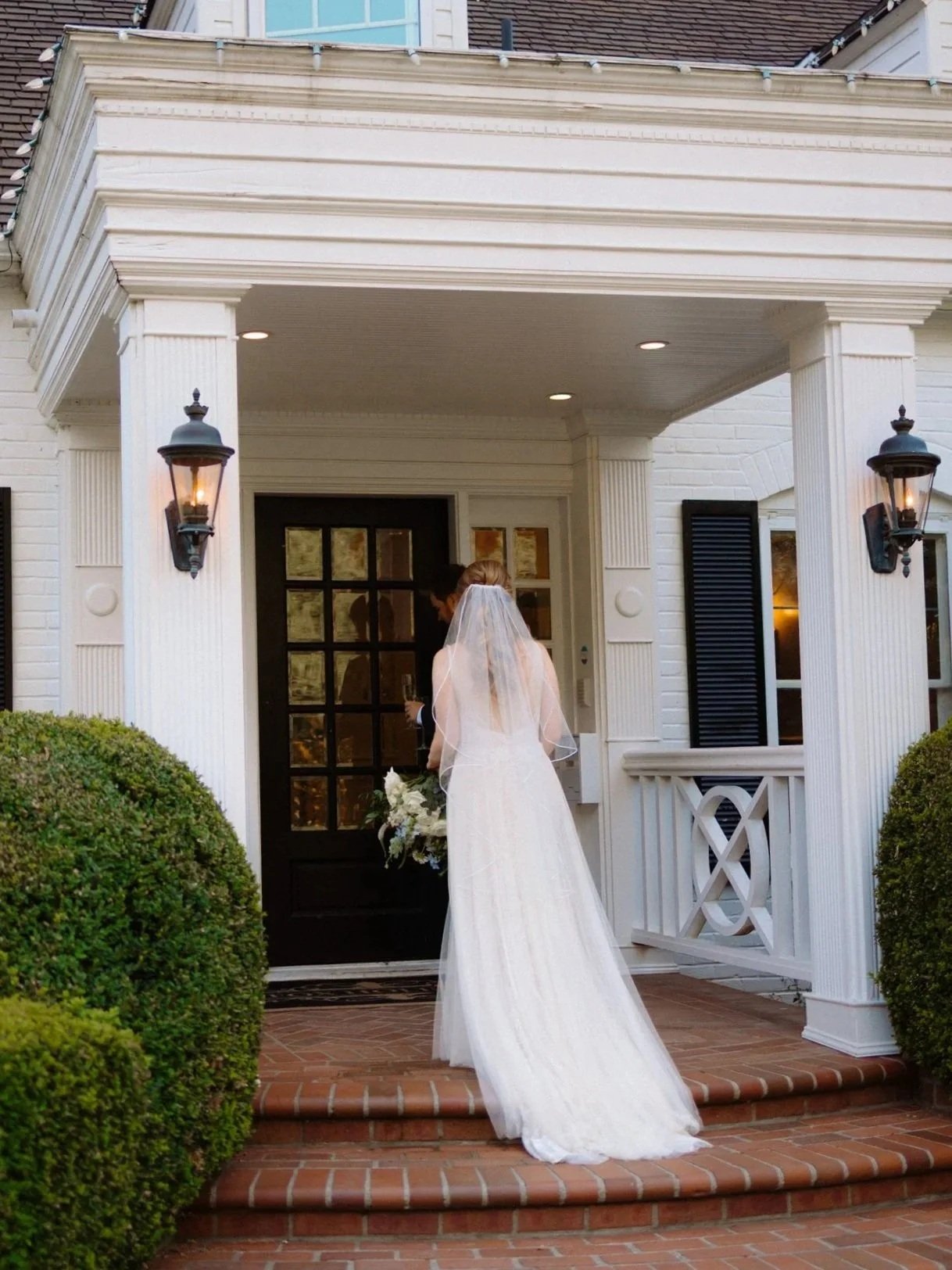 Bride in a white wedding dress with a veil, holding a bouquet of flowers, standing on steps outside a white brick house with black shutters and outdoor wall lanterns.