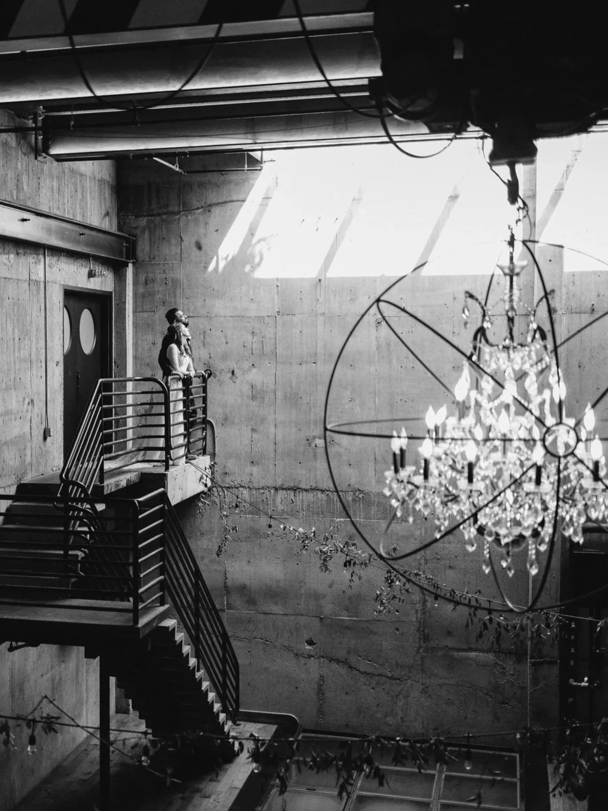 A black-and-white photo of an industrial-style interior with concrete walls, a staircase, and a chandelier hanging from the ceiling. A woman is standing on the balcony, looking out.