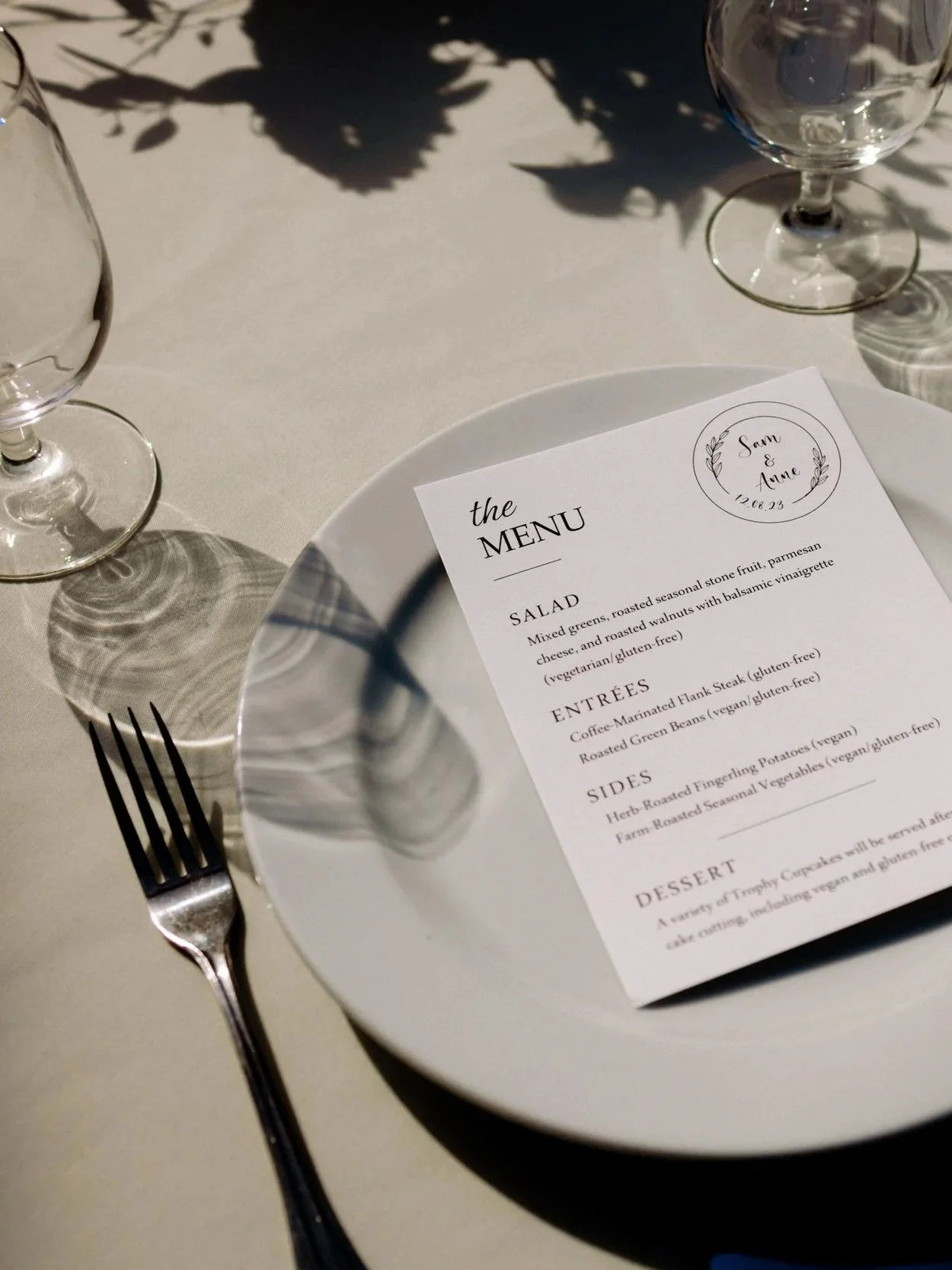 A formal table setting with a white plate, a fork on the left, a knife on the right, a water glass behind the plate, and a menu card placed on the plate. The menu lists various dishes including salads, entrées, sides, and desserts. There are shadows 