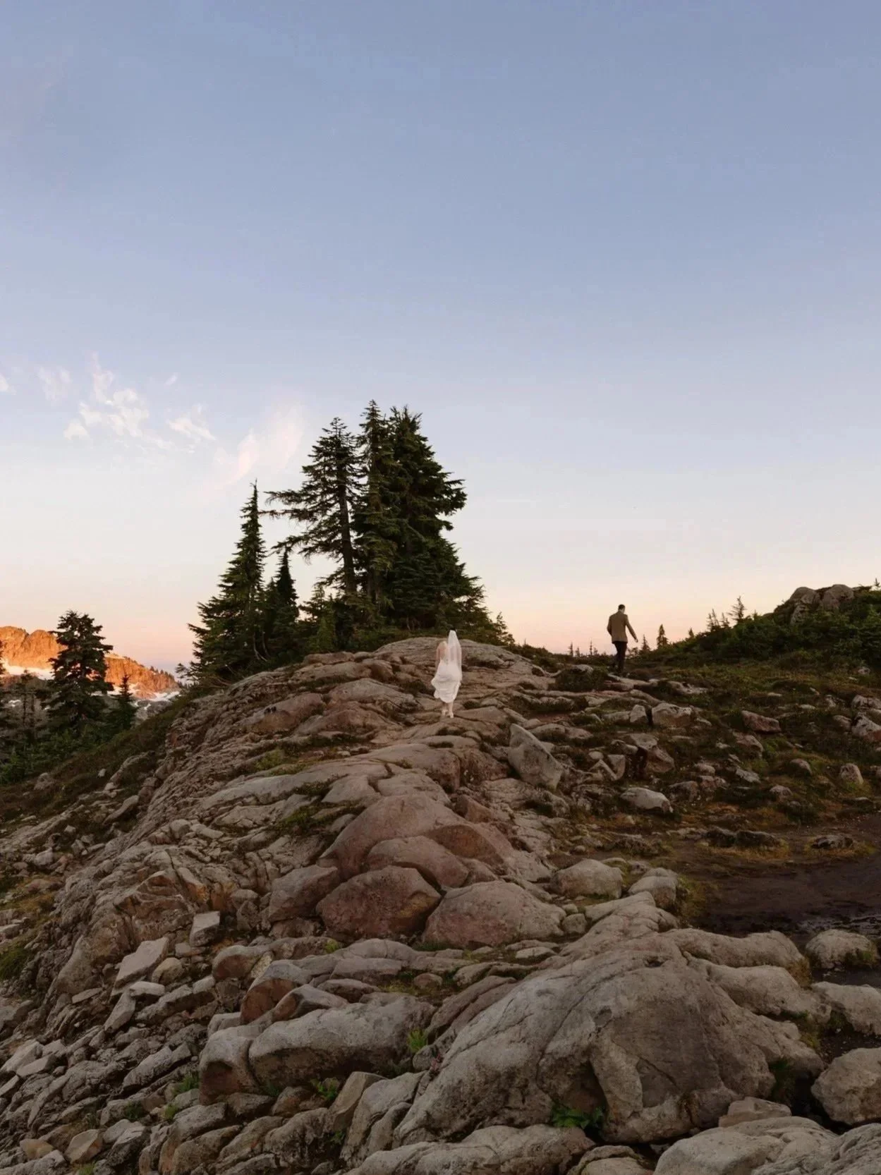 A person and a woman with a dress walking on a rocky mountain trail near a pond, with trees and snow-capped mountains in the background during sunset.