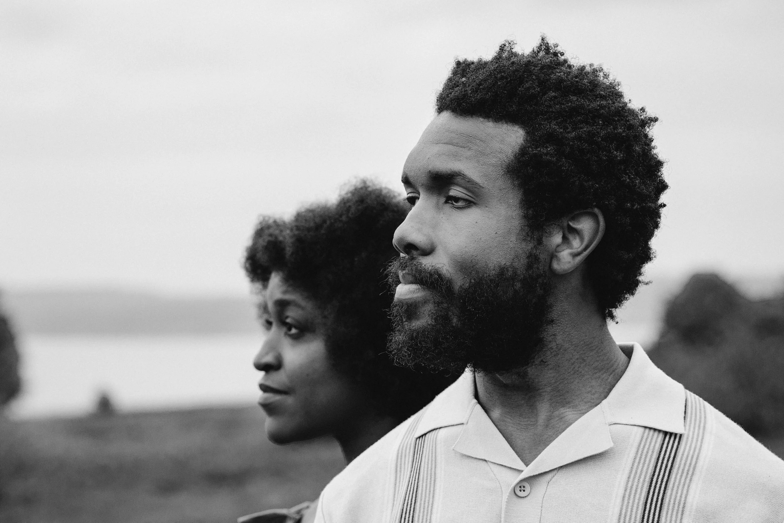 A black-and-white photo of a man and woman standing outdoors, both looking to the left with serious expressions. The man has a full beard and curly hair, wearing a collared shirt. The woman has short, curly hair.