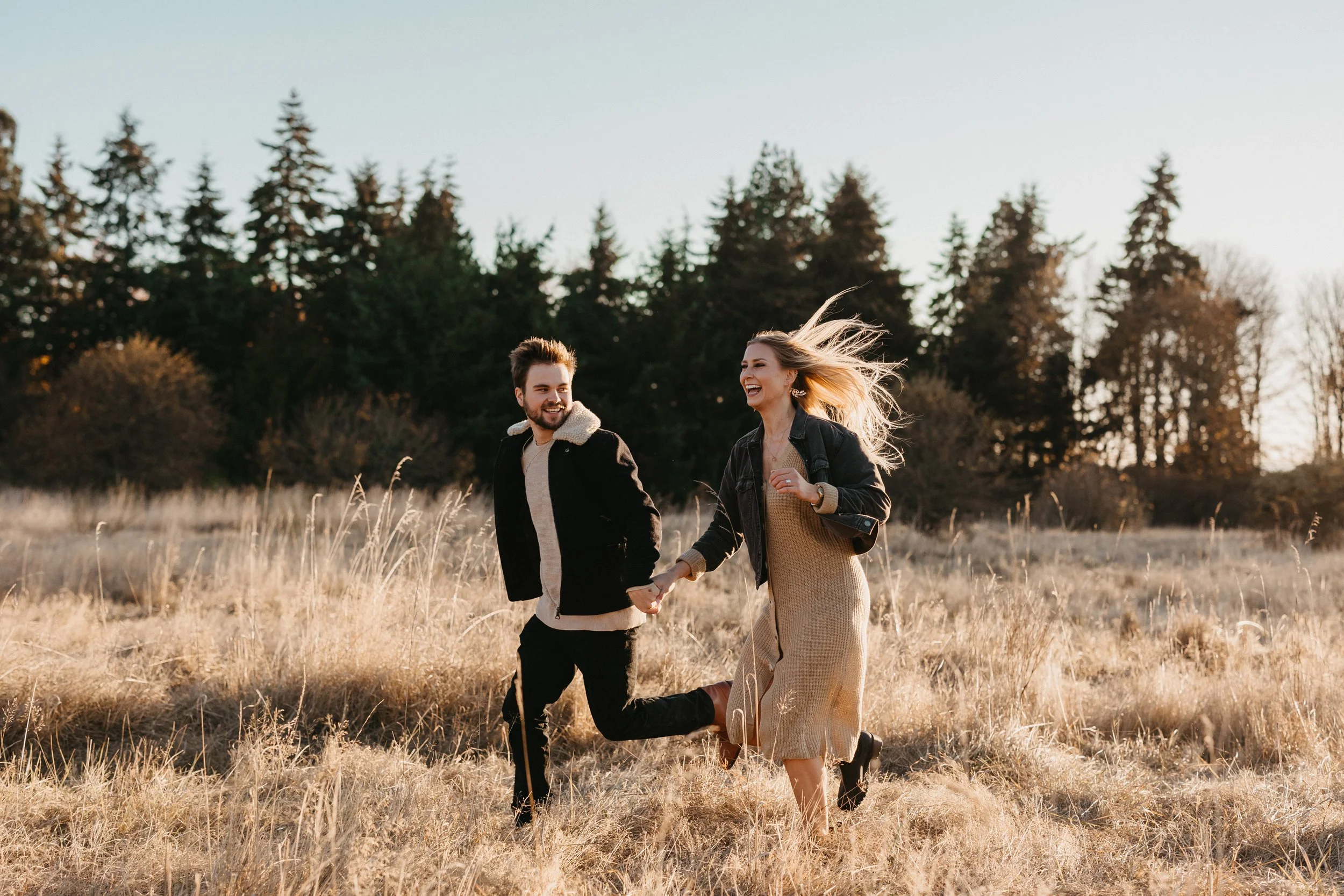A young man and woman holding hands and running through a field of tall, dry grass with trees in the background on a sunny day.