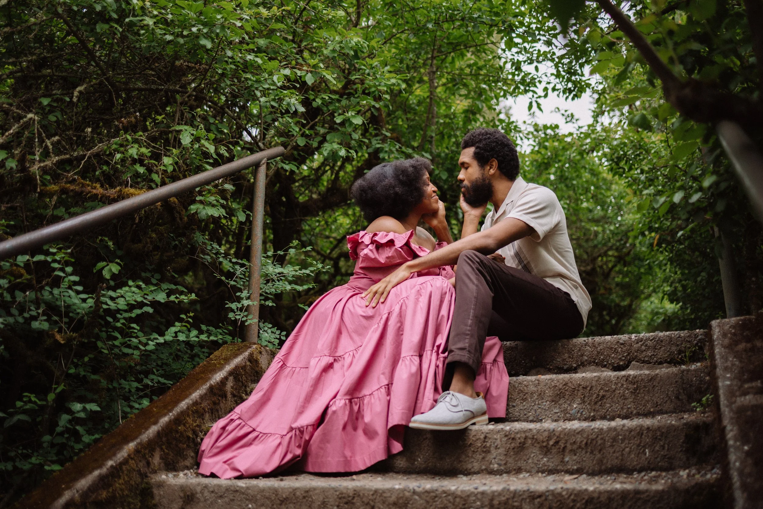 A couple sitting on outdoor concrete stairs surrounded by green foliage, gazing into each other's eyes, with the woman wearing a pink dress and the man in a light shirt and dark pants.