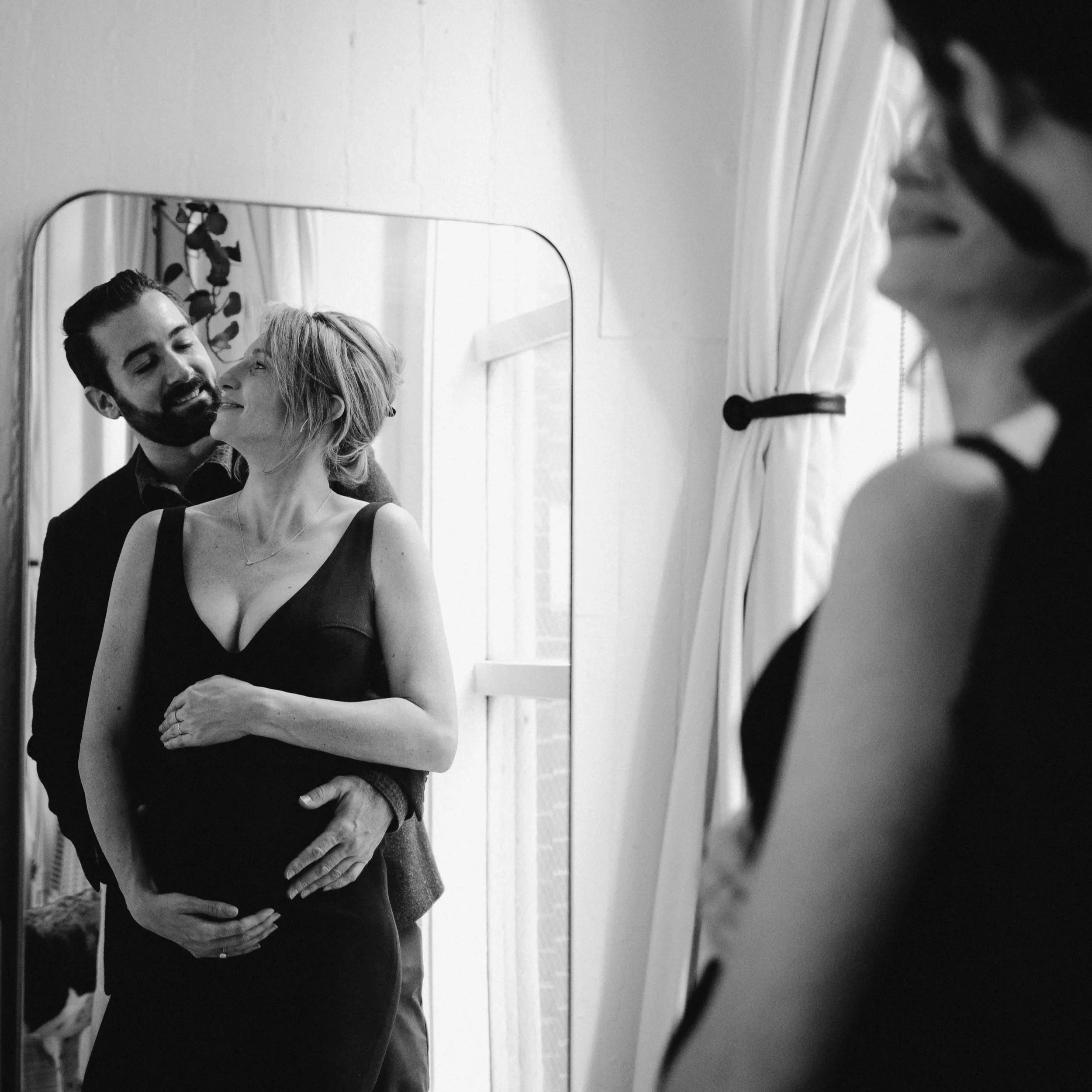 A couple looking into a mirror, with the woman holding her pregnant belly, smiling at each other, in a black-and-white photograph.