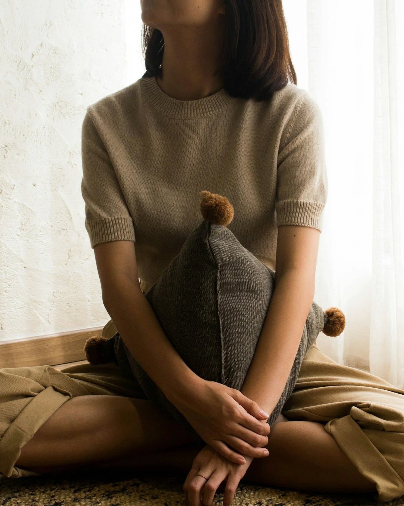 A woman sitting cross-legged, holding a gray pillow with pom poms, near a window with sheer curtains, in a softly lit room.