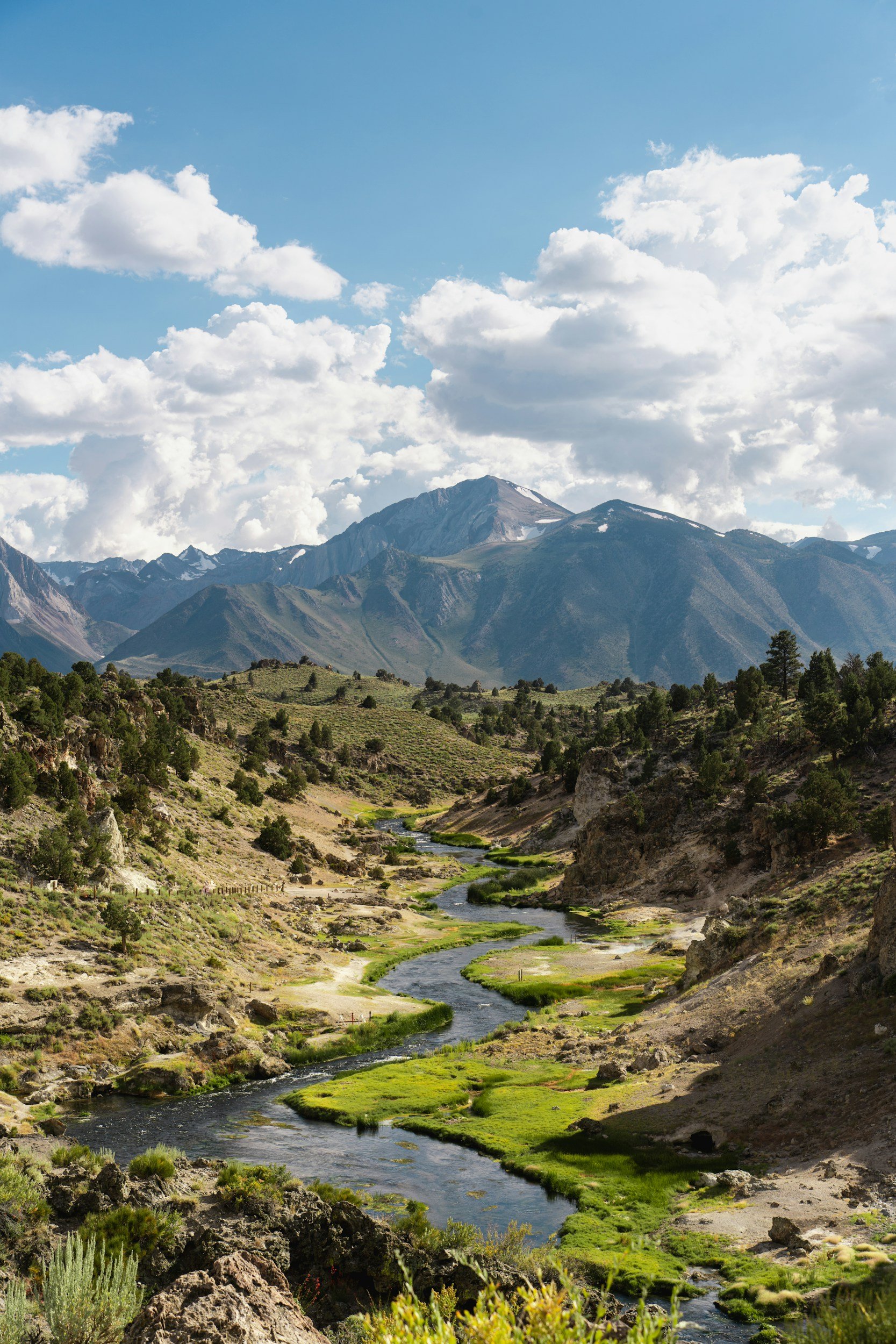 A scenic view of a winding river flowing through a mountainous landscape with green vegetation, rocky terrain, and snow-capped peaks under partly cloudy blue sky.
