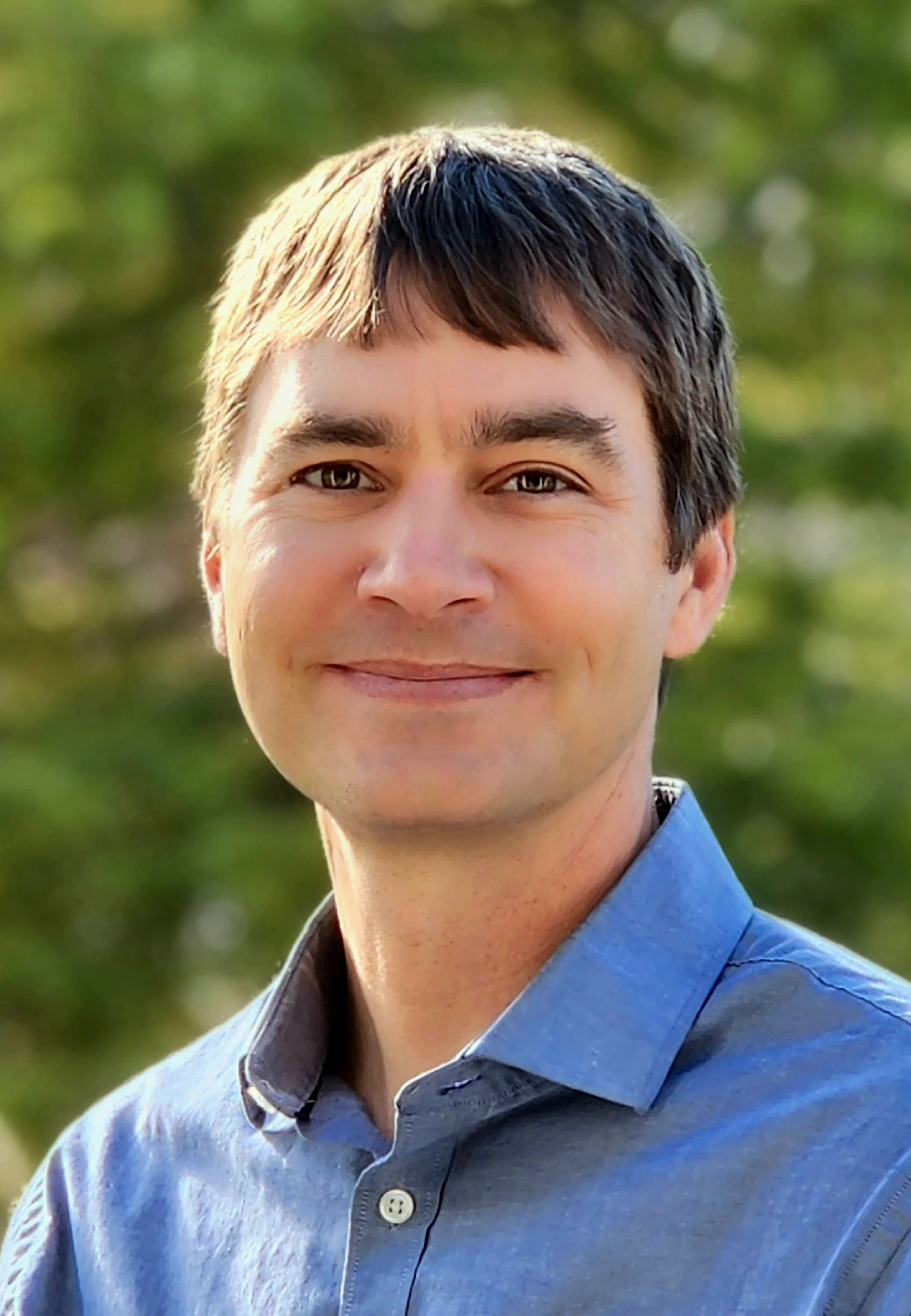 Close-up of a young man with light skin and dark brown hair, smiling outdoors with trees in the background, wearing a blue button-up shirt.