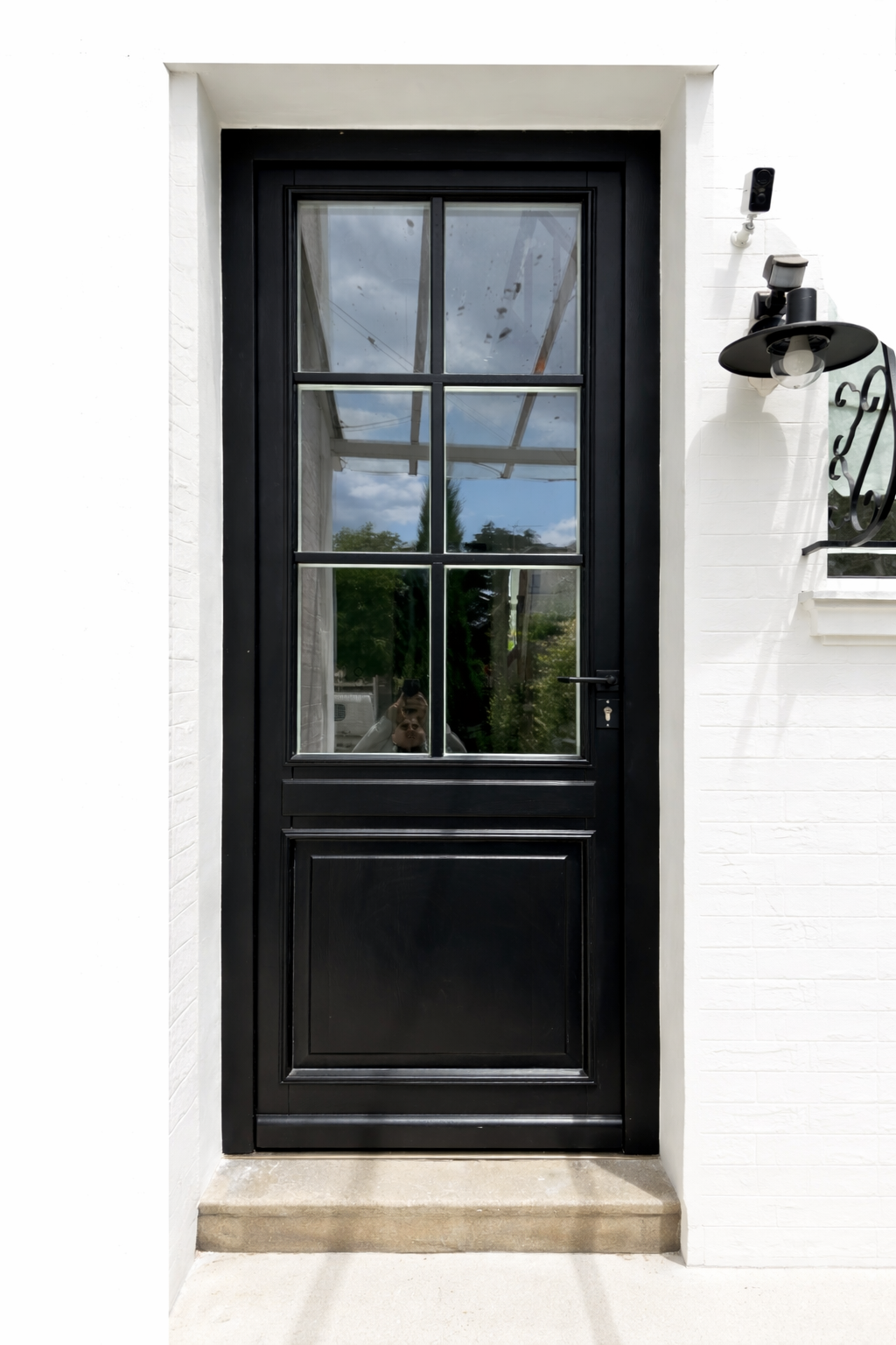 Porte noire en verre à plusieurs fenêtres, encadrée de blanc, avec une lumière murale noire à côté et une caméra de sécurité en haut à droite.