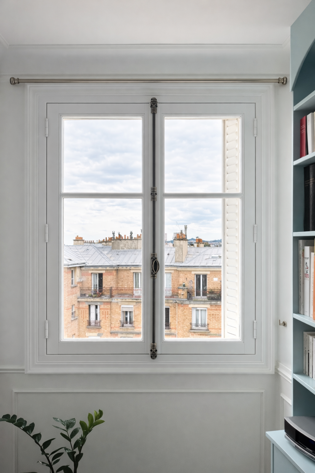 Vue vers des bâtiments résidentiels en briques avec toits en zinc, vues à travers une grande fenêtre à doubles vantaux avec cadre blanc dans une pièce lumineuse.