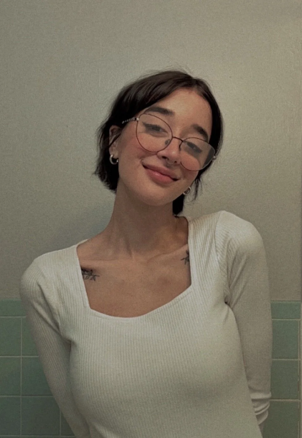 A young woman with short dark hair, wearing glasses, a white long-sleeve shirt, earrings, and tattoos on her shoulders, smiling and posing in front of a plain wall.