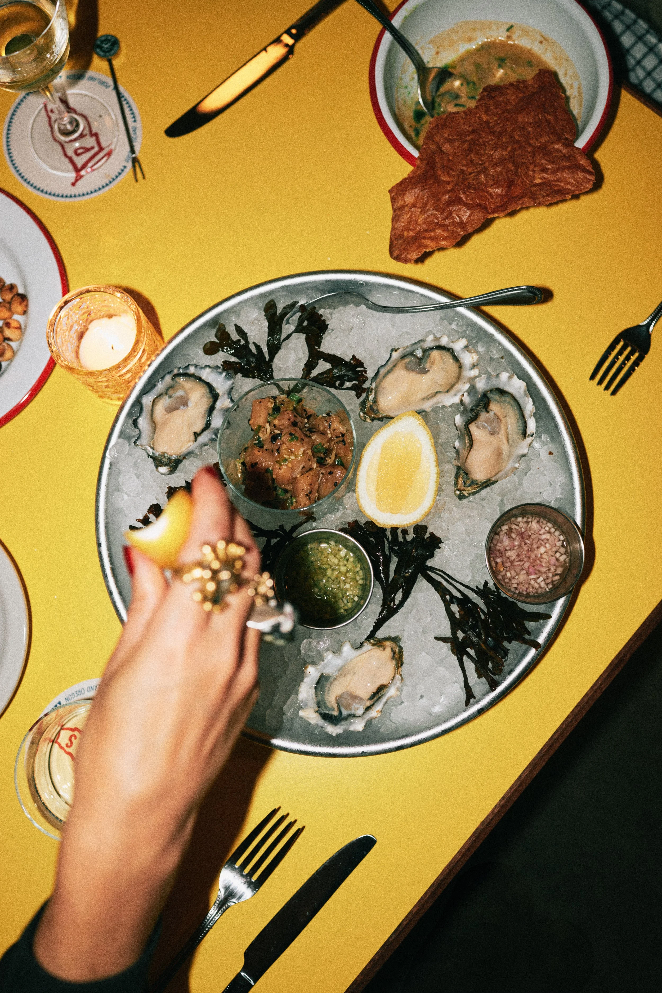 A round platter of raw oysters on ice with lemon wedges, sauce, and garnishes, at a dinner table with a yellow tablecloth, surrounded by plates, wine glasses, and cutlery.