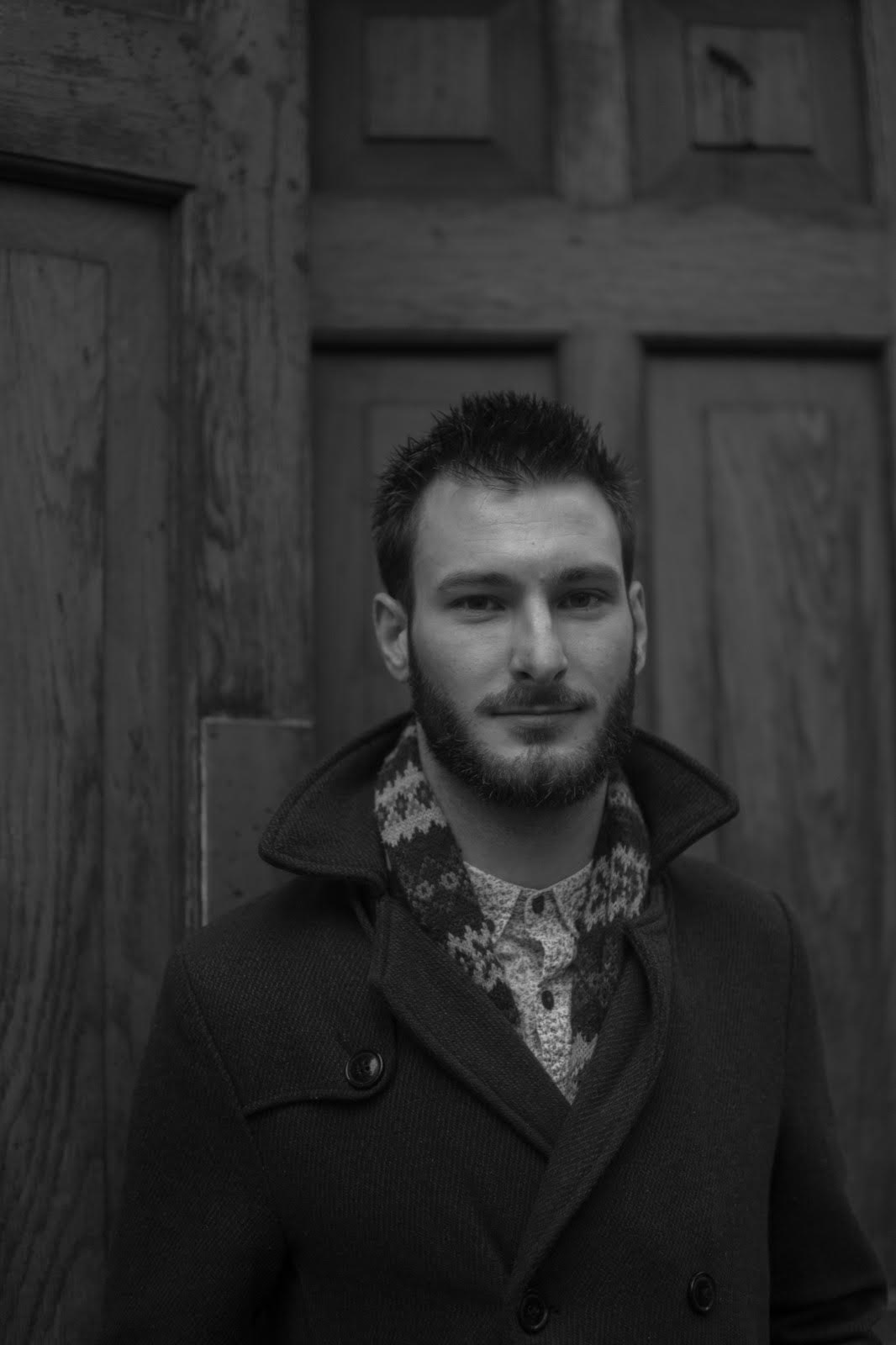 A young man with a beard and short hair standing in front of a wooden door, wearing a dark coat and a patterned shirt.