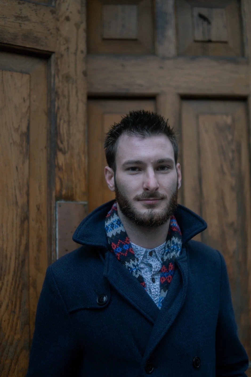 A young man with short dark hair and a beard standing in front of a wooden wall or door, wearing a navy coat and a patterned shirt underneath.