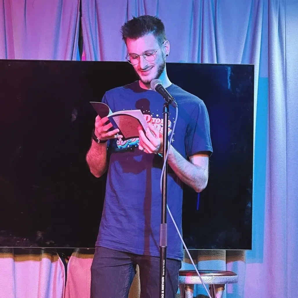 A young man with glasses, short styled hair, and a beard, standing on stage reading from a small book or booklet into a microphone on a stand, with a large TV or monitor behind him and purple curtains to the side. He's smiling and wearing a blue T-shirt.