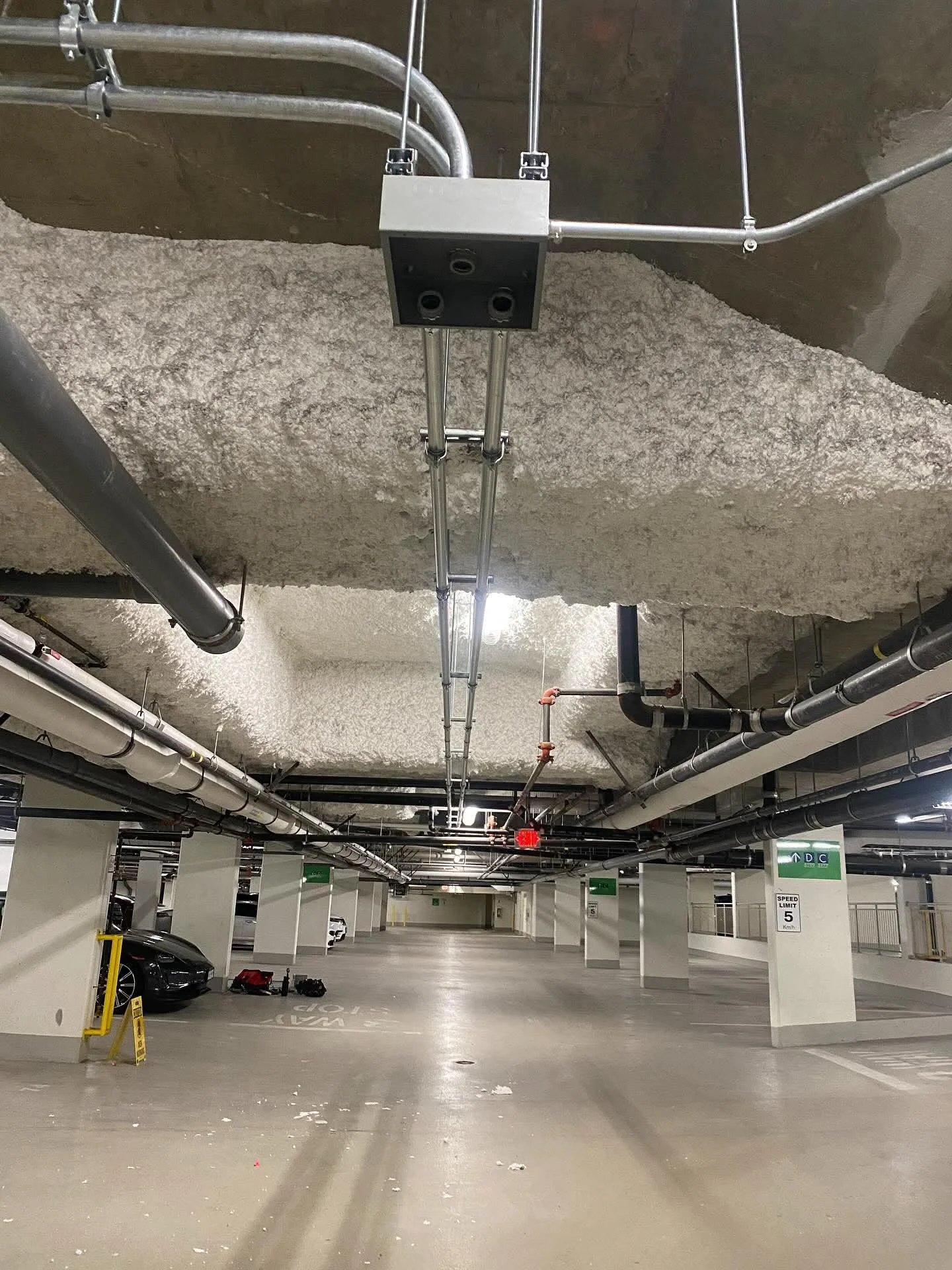 Underground parking garage with parked cars, visible pipes, and sprinkler system on the ceiling.