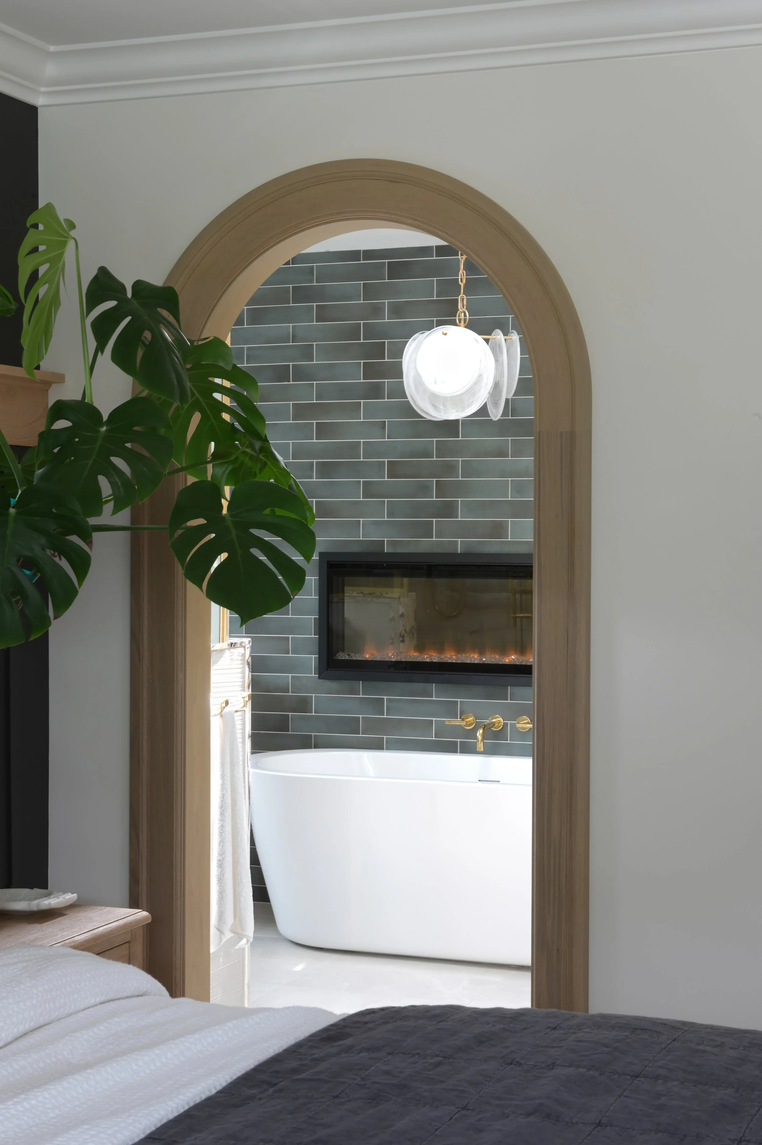 A bedroom with a partial view of a bathroom through an arched doorway. The bathroom features a white bathtub, gold fixtures, a dark gray tiled wall, and a mounted fireplace. A large green plant is in the foreground.