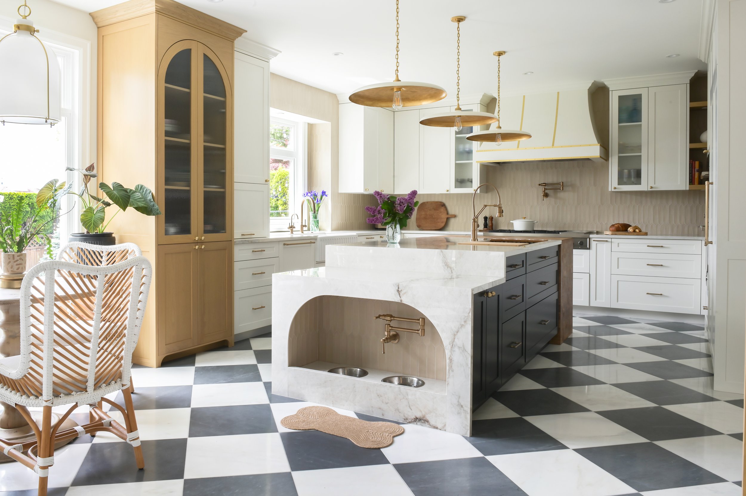 Bright, modern kitchen with black and white checkered floor, white cabinets, a central marble island with a built-in pet water and food area, gold pendant lighting, and various plants and flowers.