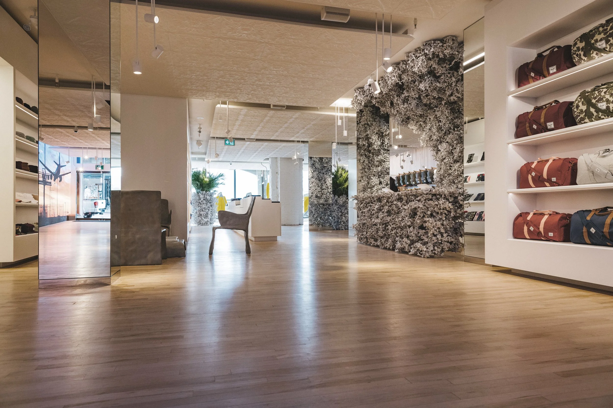 Interior of a retail store with wooden flooring, chairs, and display shelves filled with bags and accessories. There are decorative elements like a textured black and white wall feature, plants, and modern lighting.