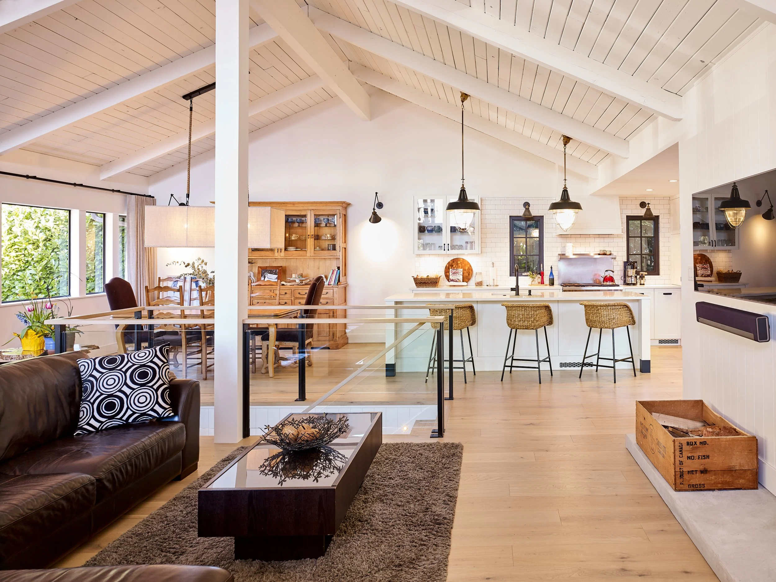 Open-concept living and kitchen area with vaulted white ceiling, wooden flooring, and large windows. The living room has a brown leather couch and a dark coffee table with a decorative bowl. The kitchen features white cabinets, a long island with fou
