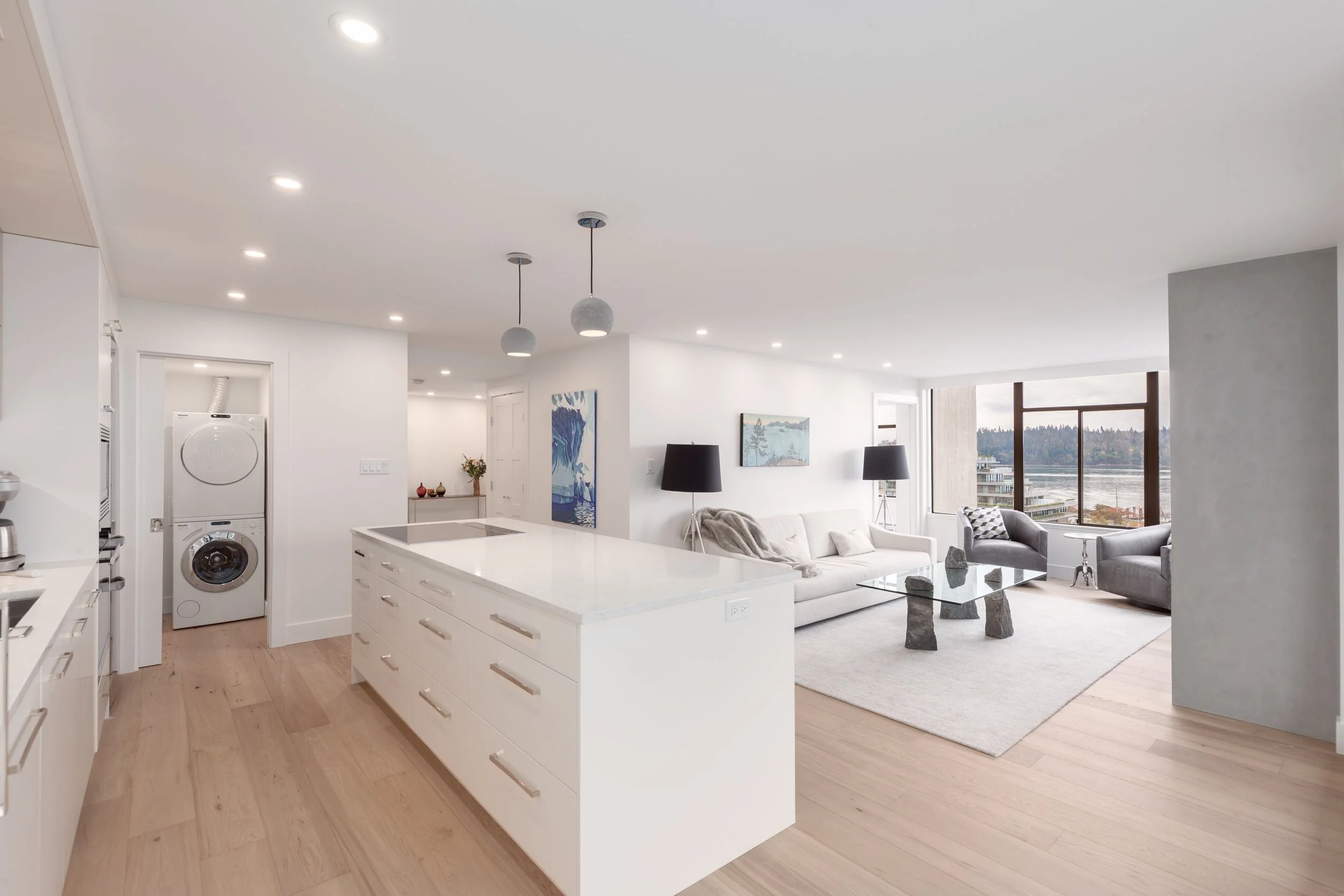 Modern open-concept living room and kitchen with white cabinets, light wood flooring, a large window with a view of water and trees, and minimal decor.