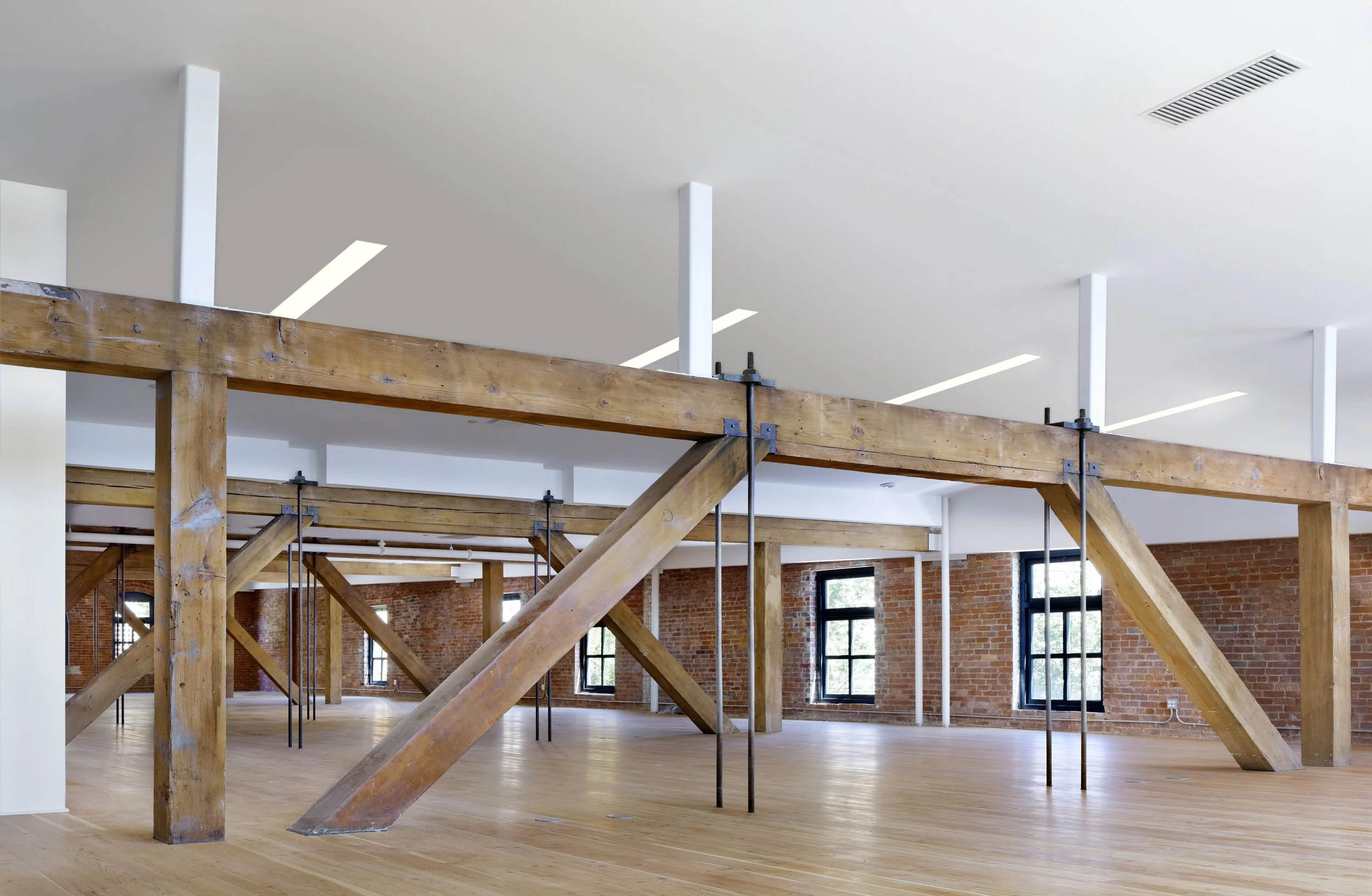Empty industrial-style interior space with exposed brick walls, large windows, and wooden support beams.