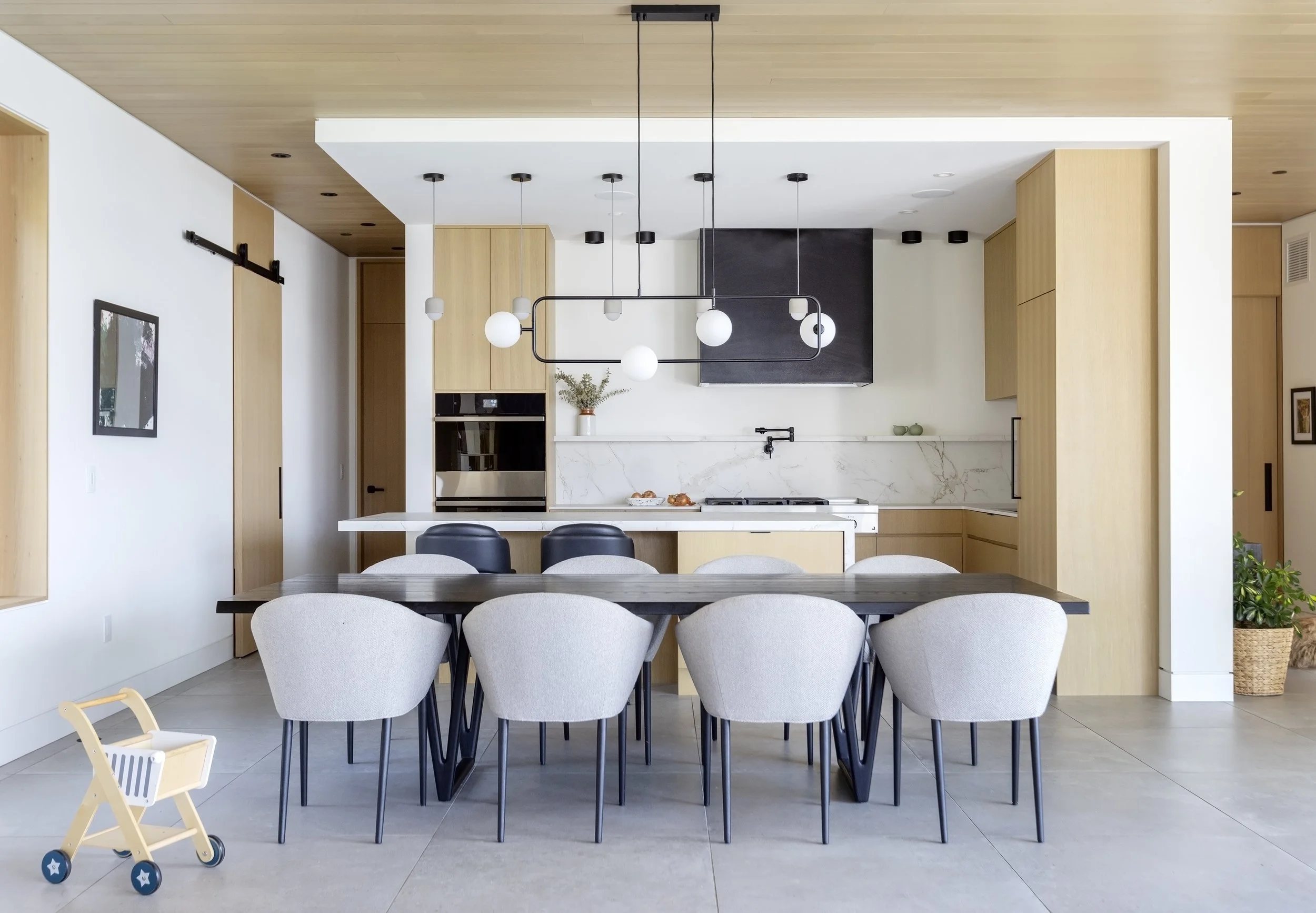 Modern minimalist kitchen and dining area with light wood cabinets, white marble backsplash, black chairs, off-white upholstered dining chairs, and a black ceiling light fixture.
