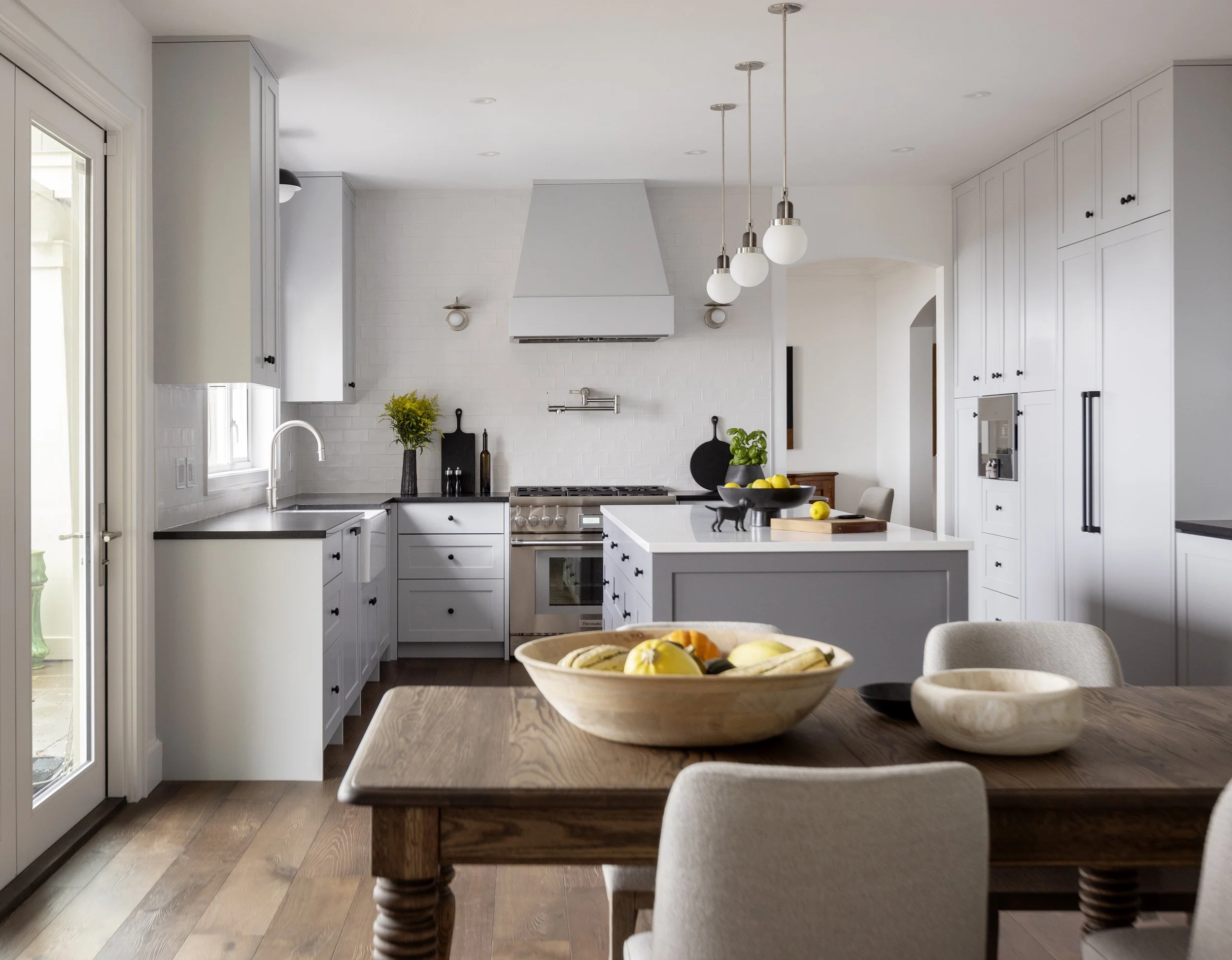 Modern white kitchen with wooden dining table, fruit bowls, pendant lighting, and stainless steel appliances.