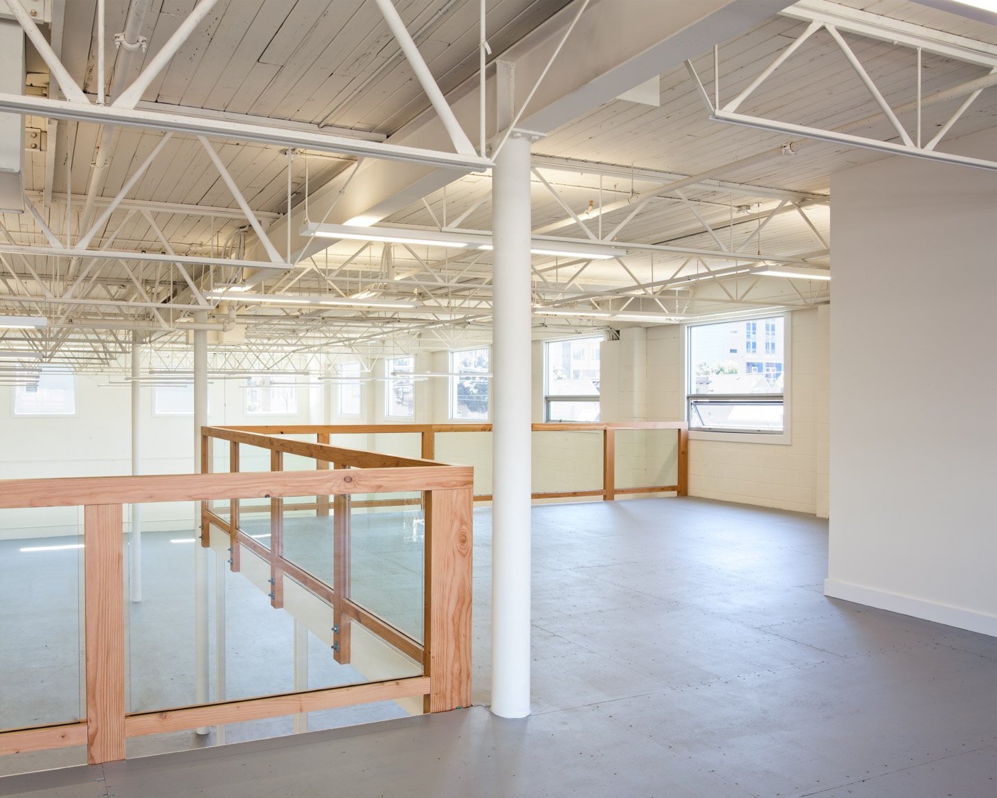 Empty modern interior with white walls, large windows, exposed ceiling beams, and a wooden railing around an upper level.