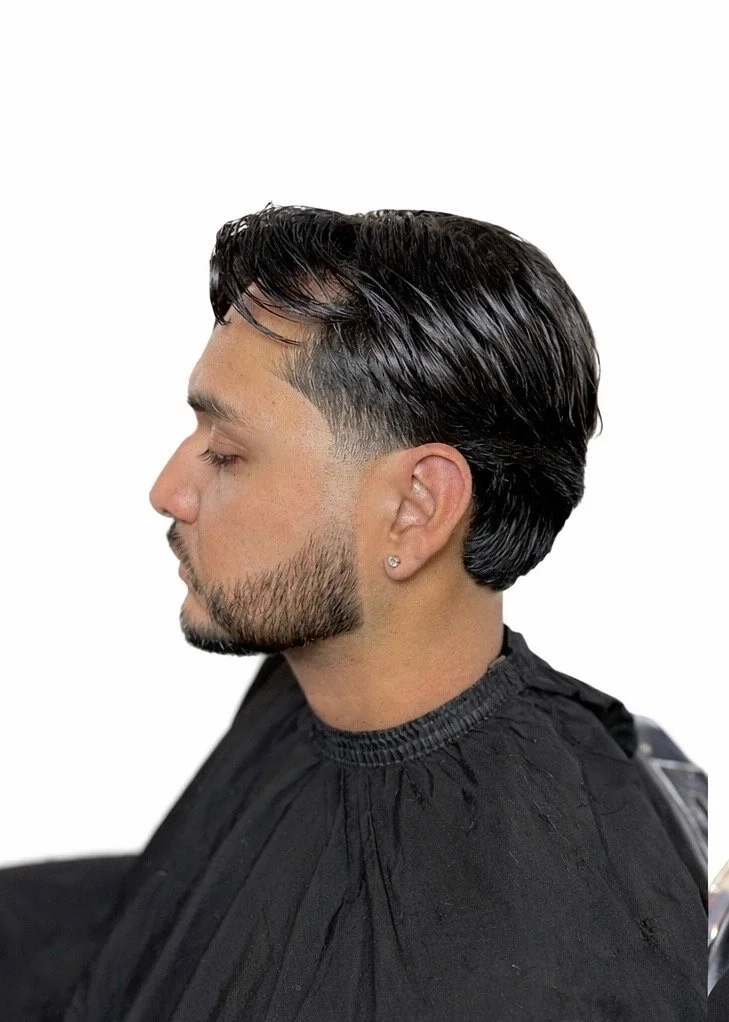 Man with dark, sleek, and styled hair with a faded undercut, beard, and earring in ear, sitting in a black barber cape against a white background.