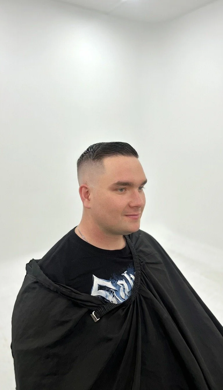 A man sitting in a salon chair with a black cape, after getting a haircut with a fade hairstyle, in front of plain white walls.