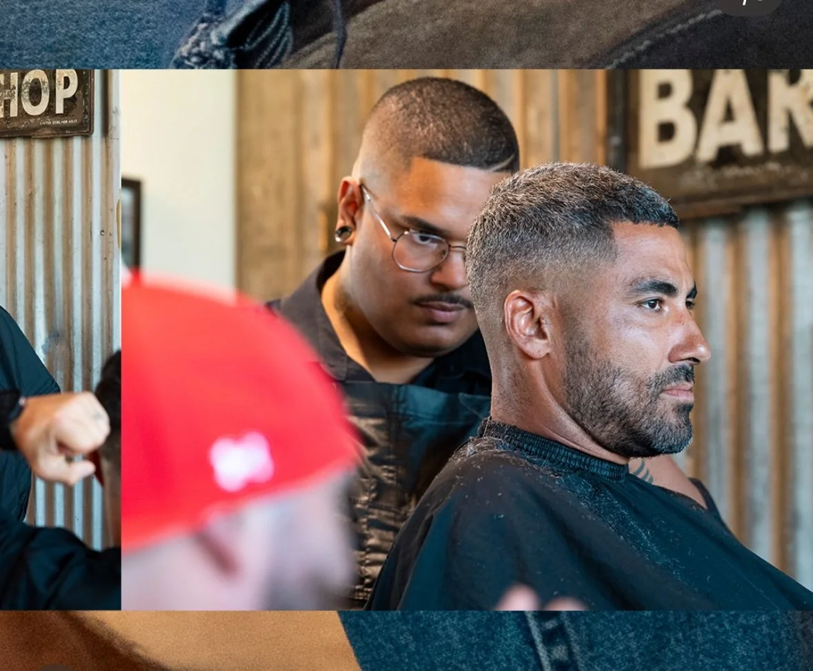 A man with a neat beard and short gray hair getting a haircut at a barbershop, with a young male barber closely examining him.
