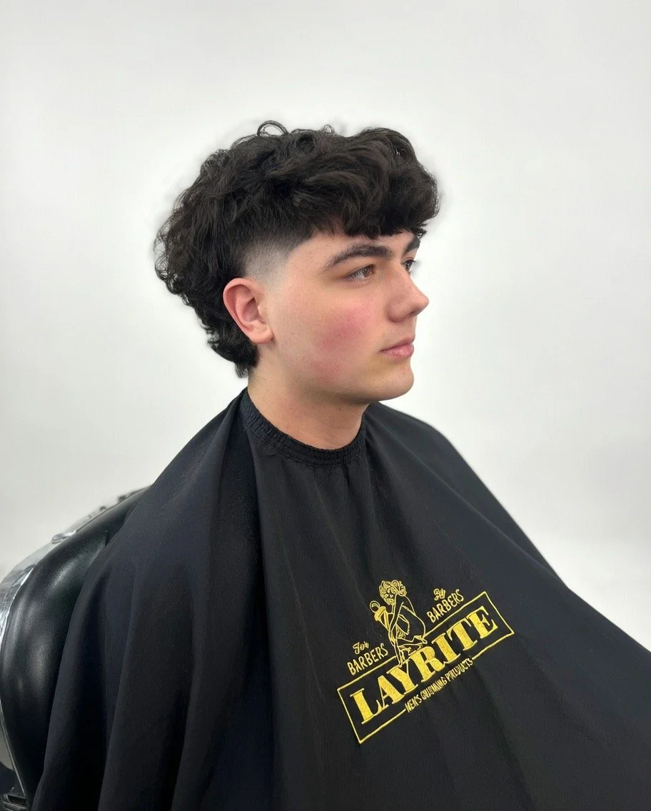 Young man with dark, curly hair and a skin fade haircut sitting in a salon chair, wearing a black cape with yellow text that reads 'LAVRTE: Men Barbers, Barbers, Men's Grooming Products'.