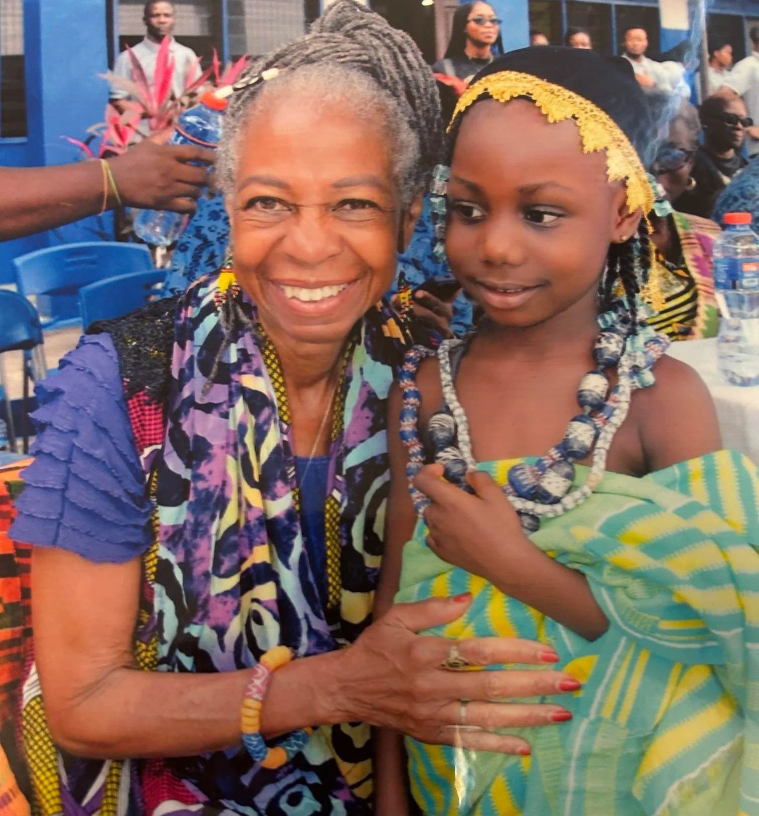 An elderly woman and a young girl wearing traditional African clothing at an outdoor event, smiling and posing for the camera.