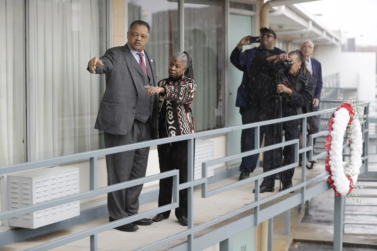 A group of people on a balcony, with two men and one woman in the foreground, possibly during a memorial or special event. Some individuals are taking photos or videos.