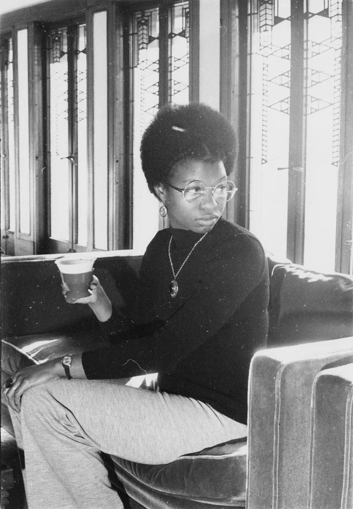 A woman with short, curly hair, wearing glasses, earrings, a necklace, and a long-sleeve black top. She is sitting on a bench by large windows with decorative grids, holding a paper cup, and looking to her right.