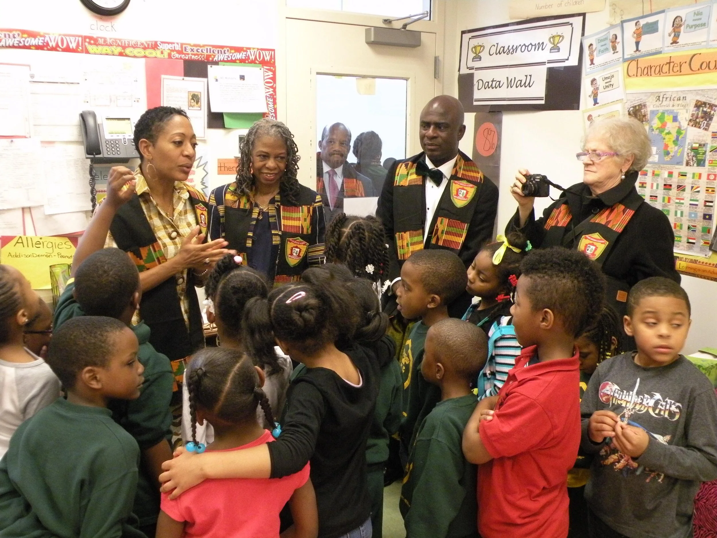 Adult educators or guest speakers, dressed in culturally significant attire with African patterns, engaging with elementary school students in a classroom setting. Students are gathered around them, listening attentively, with classroom decorations visible in the background.