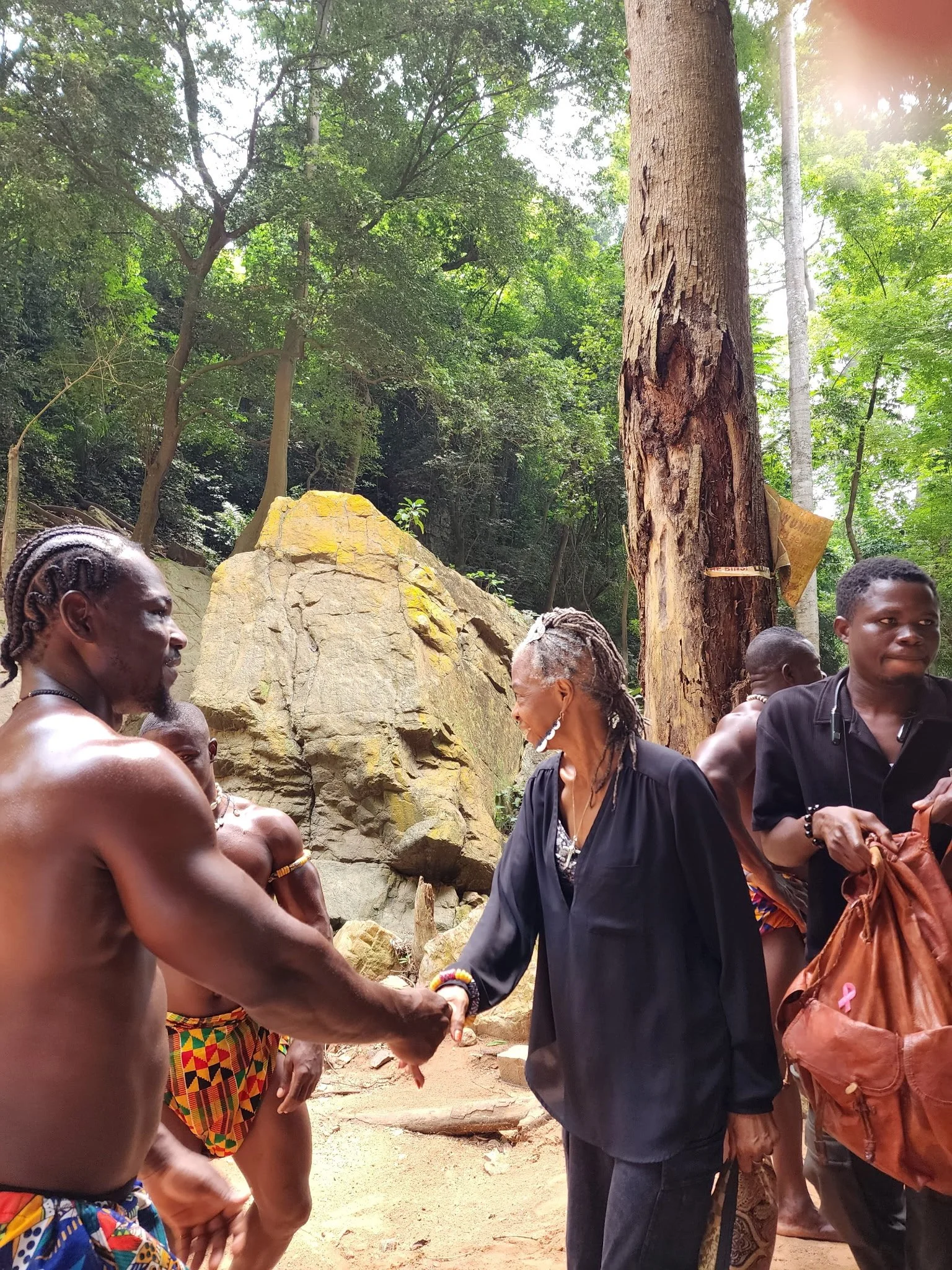 A woman with gray hair shaking hands with a shirtless man in a colorful loincloth in a forested area.