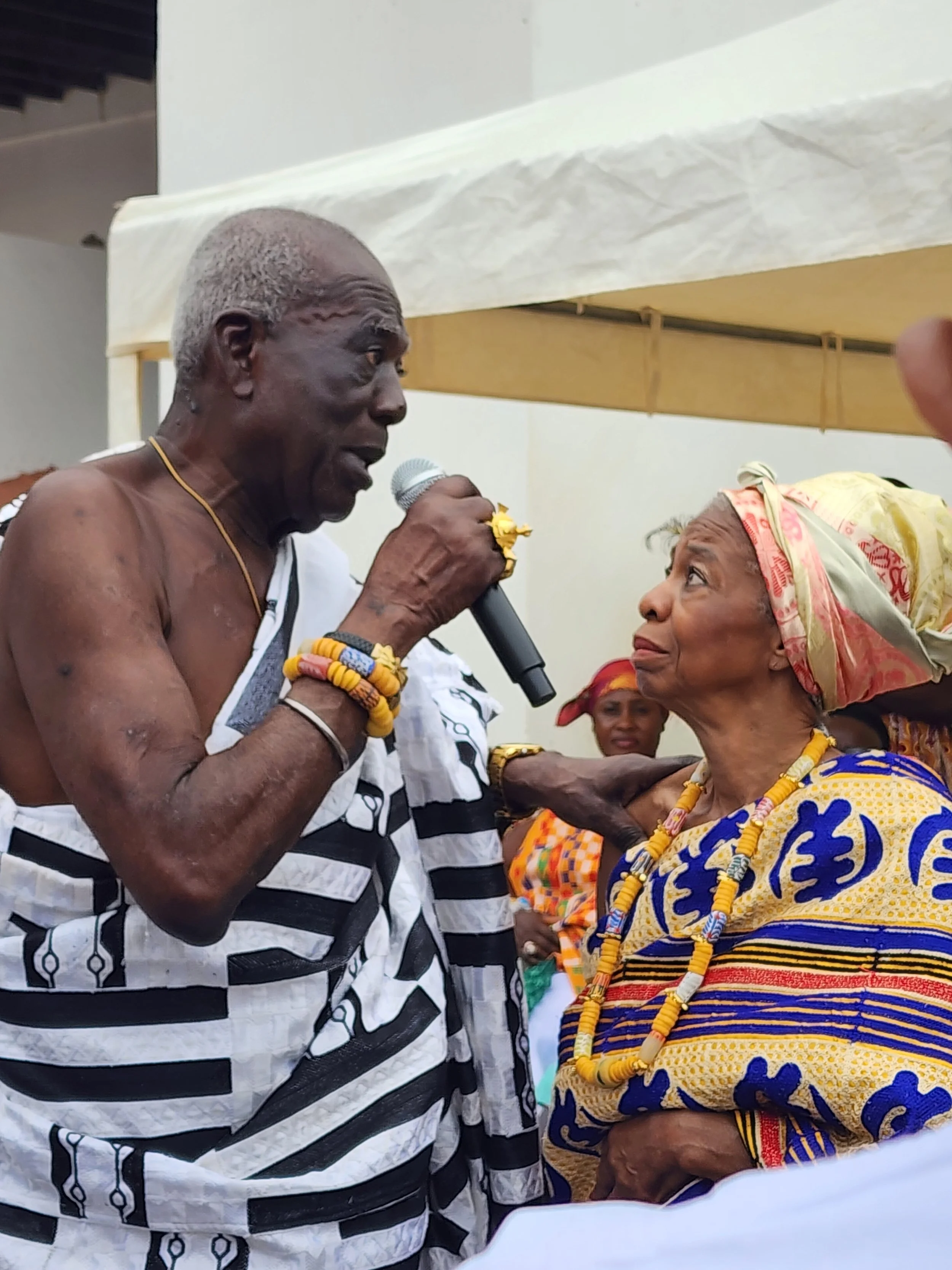 An elderly man dressed in traditional African attire speaking into a microphone, with an elderly woman dressed in colorful traditional clothing and headscarf looking at him, in a cultural event setting.