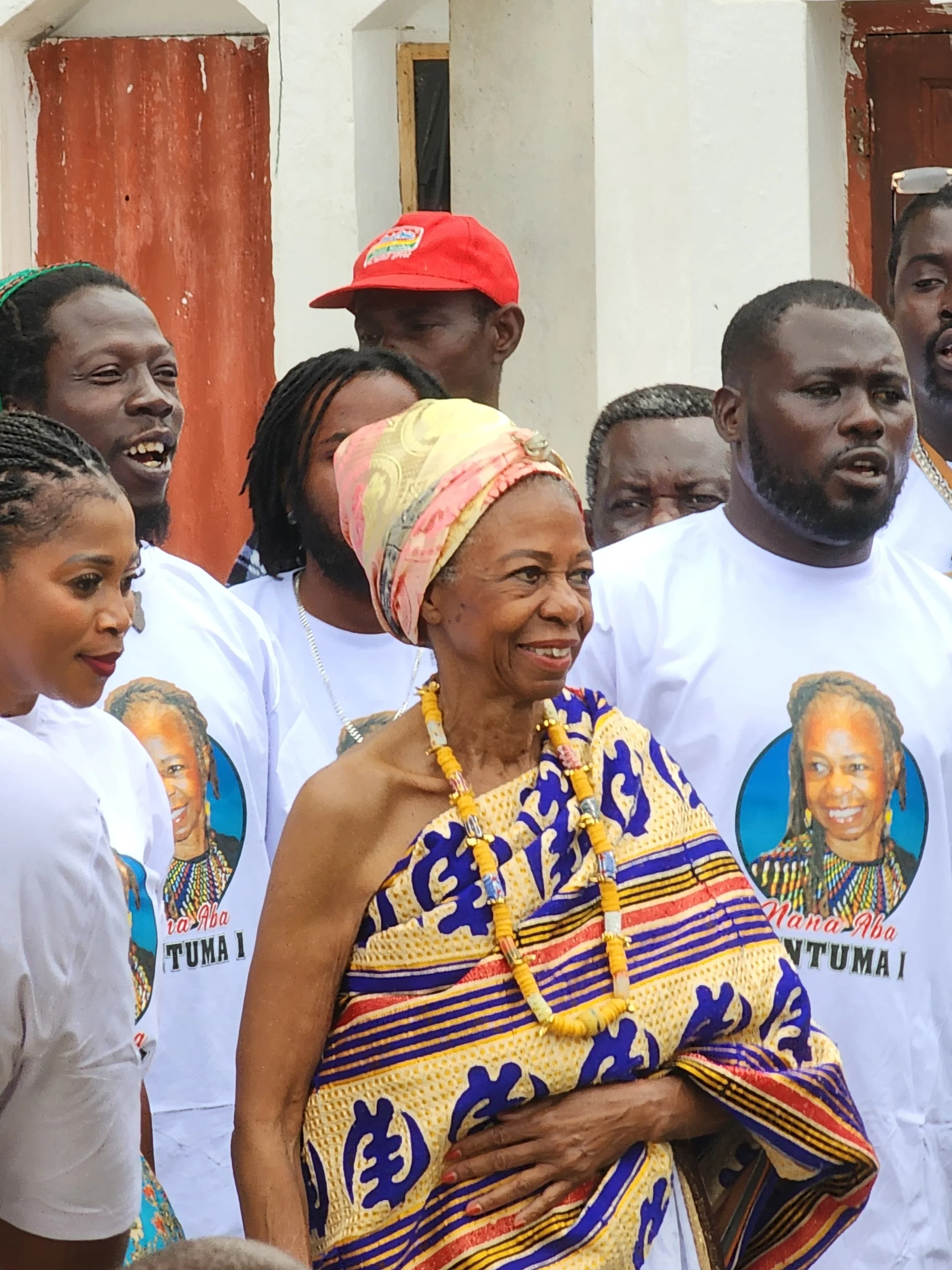 A group of people, including an elderly woman dressed in traditional African clothing and accessories, gathered outdoors during a celebration or gathering.